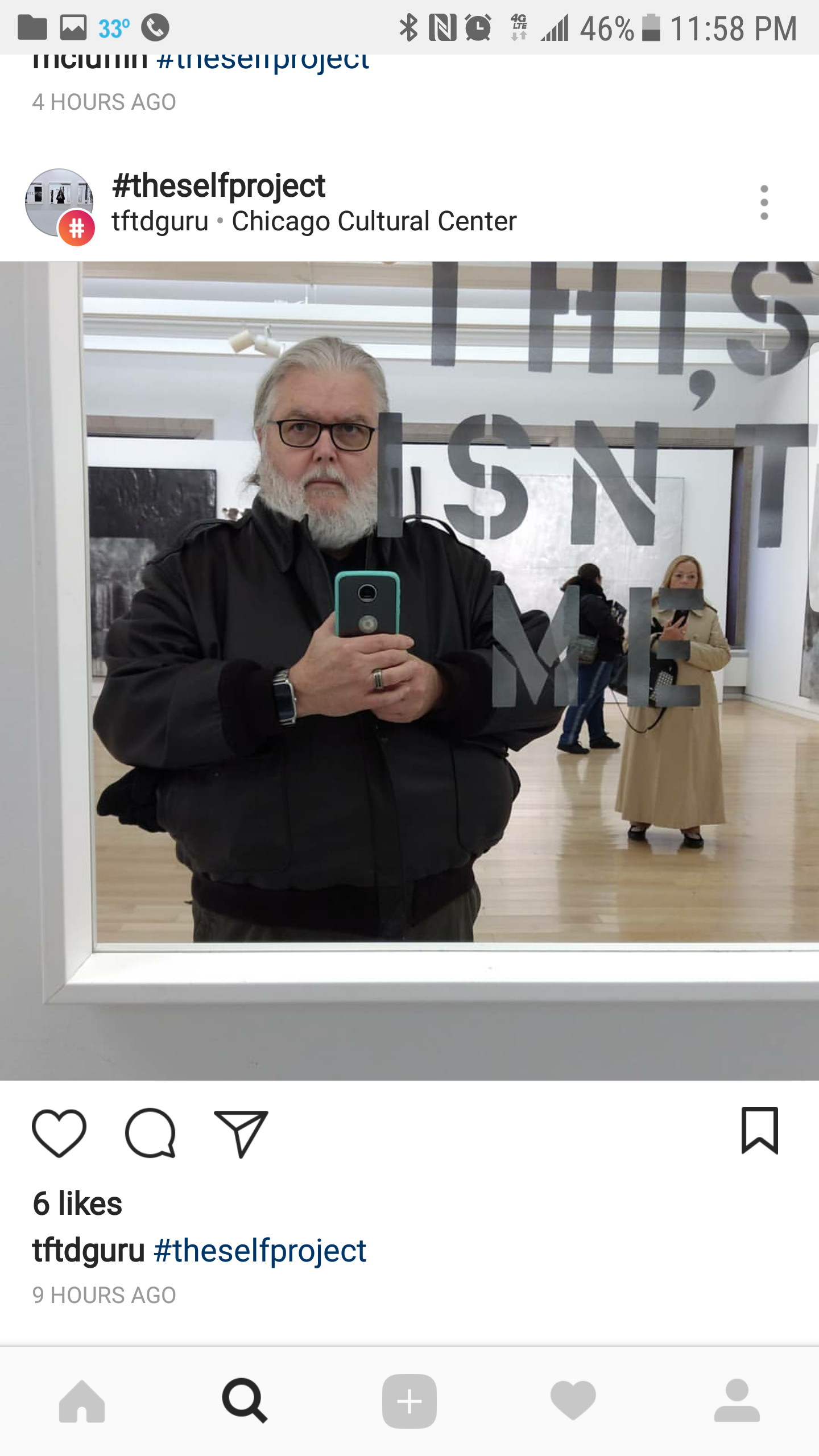 A man with gray hair and glasses taking a selfie in a mirror at an art gallery, with a woman in a beige coat and other visitors in the background.