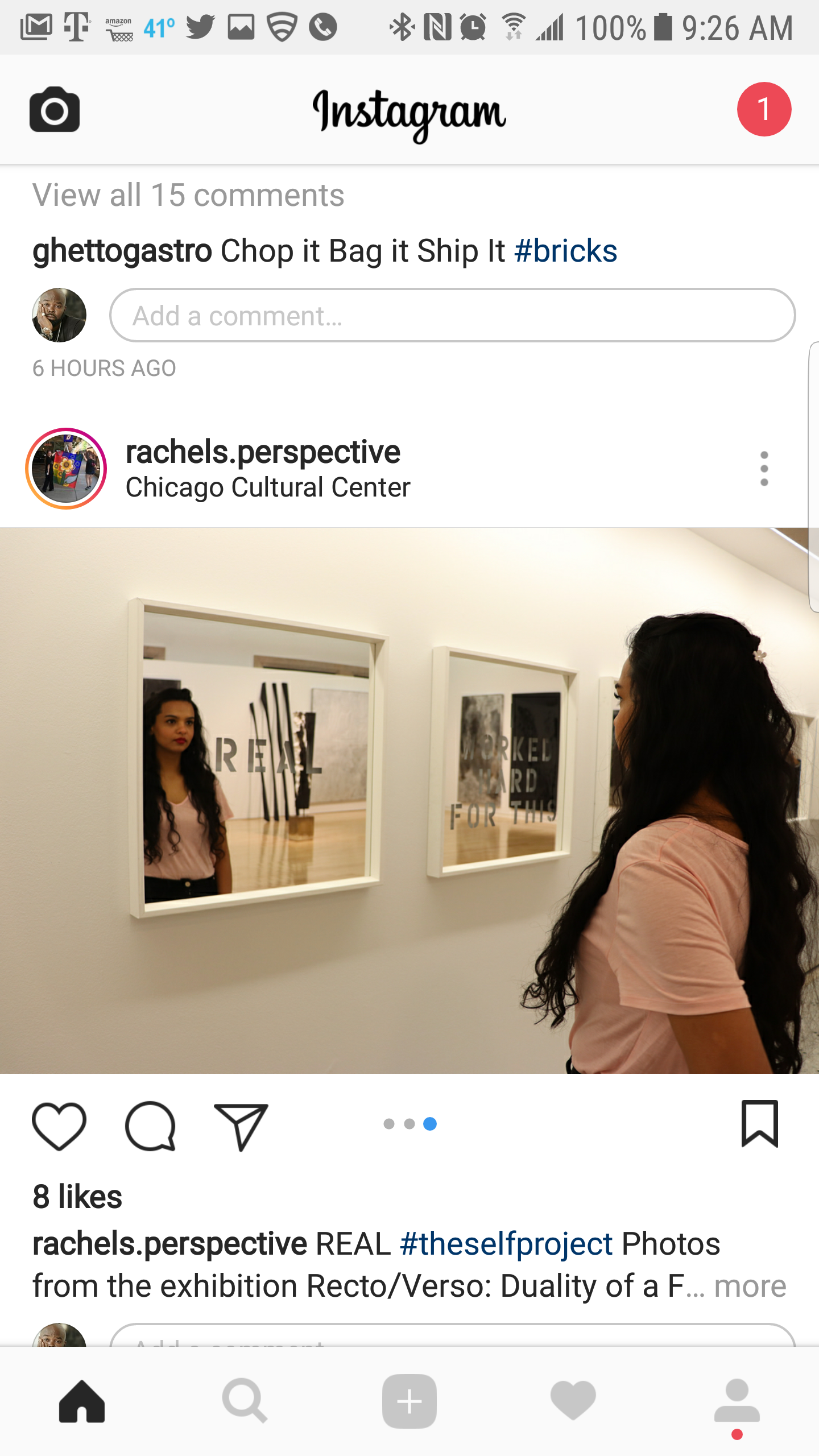 A woman with long dark hair wearing a pink shirt is looking at a mirror in an art gallery. The mirror reflects her face and part of her upper body, along with a background of artworks on the gallery wall.