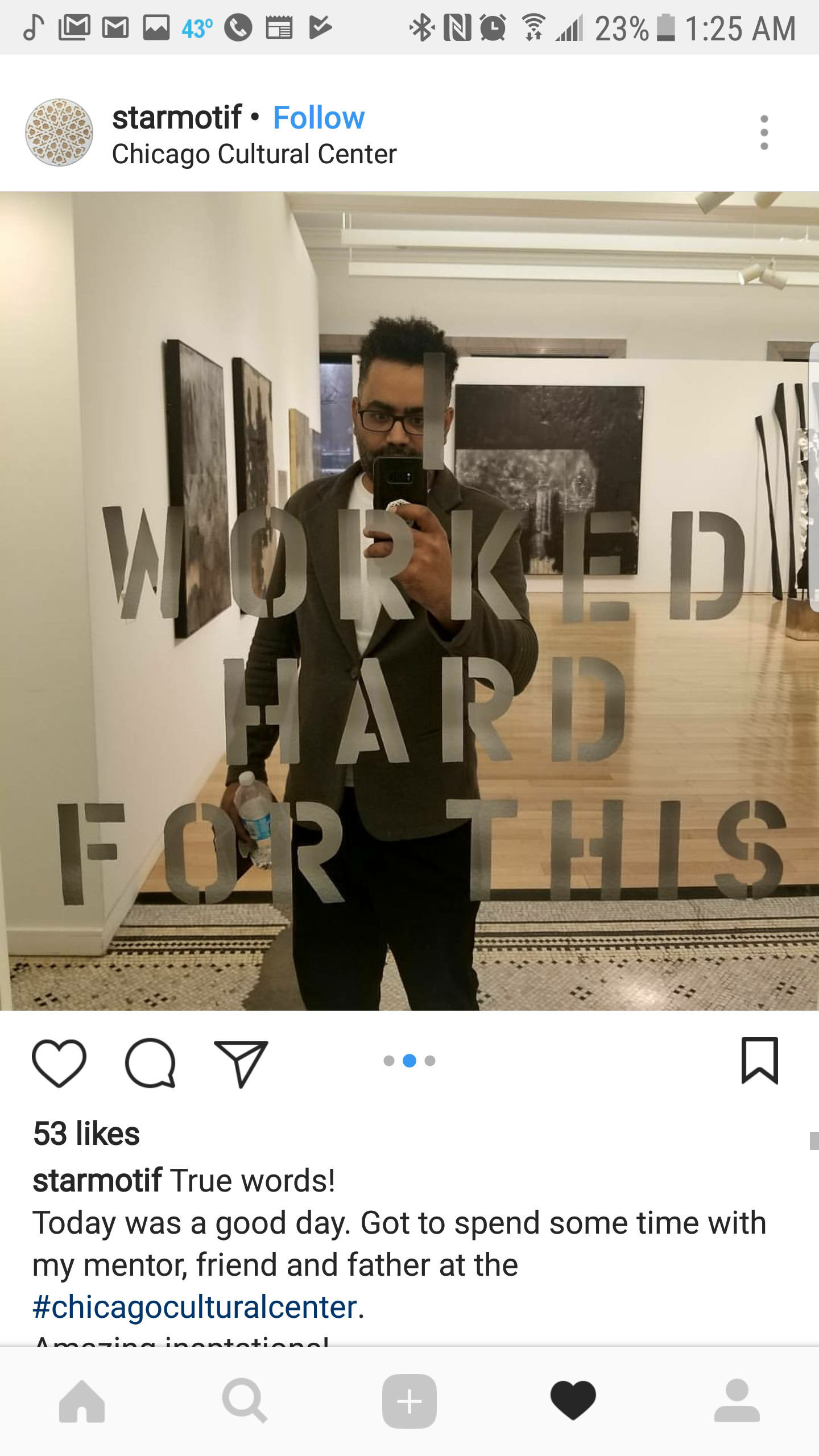 A man taking a mirror selfie at the Chicago Cultural Center inside an art gallery. The mirror has the words "WORKED HARD FOR THIS" written on it. The man is holding a water bottle and is dressed in a dark blazer with a white shirt.