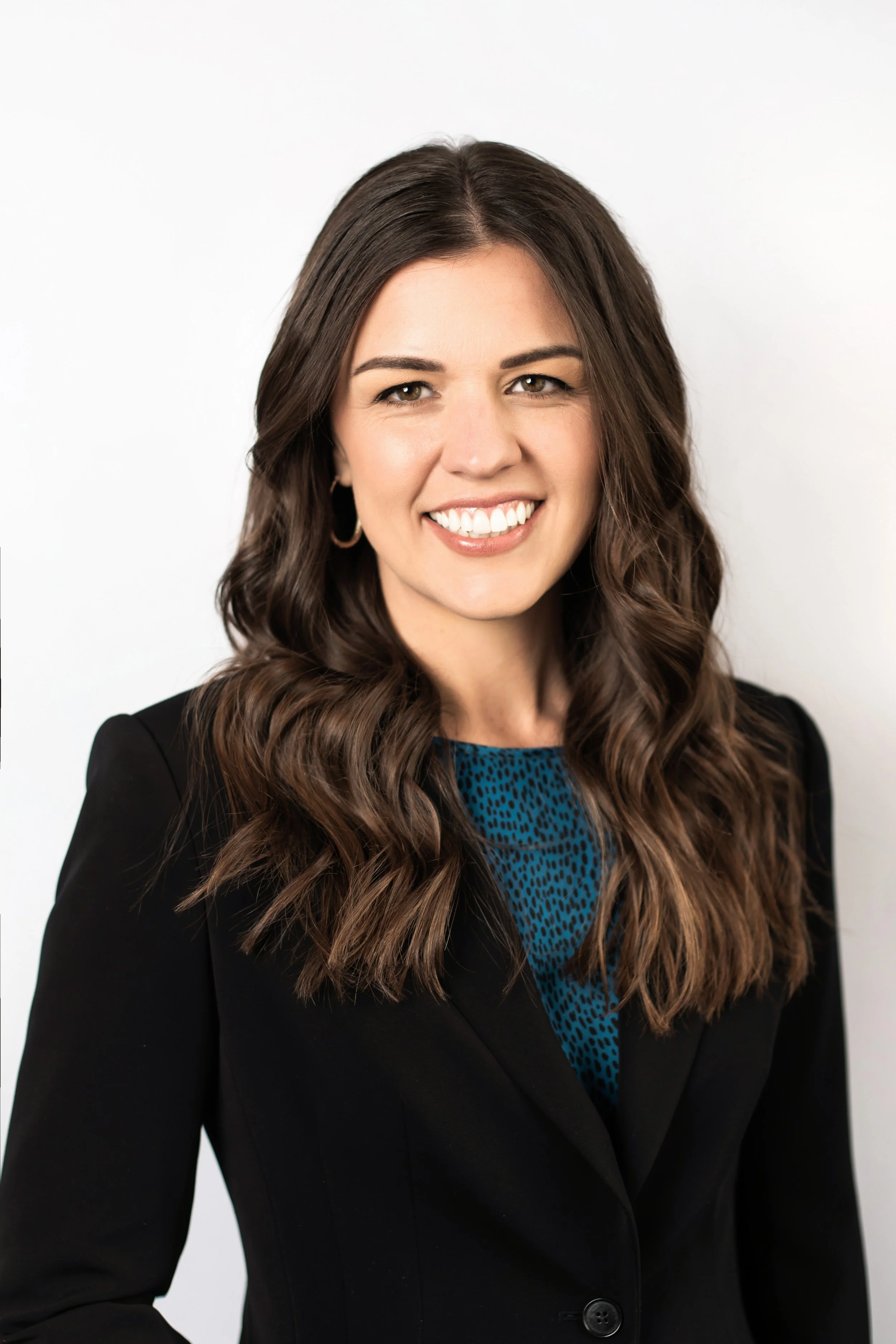 Headshot of a woman with long wavy brown hair wearing a black blazer and blue patterned blouse, smiling against a white background.