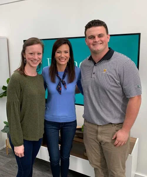 Three people standing together indoors in front of a large screen, smiling at the camera.