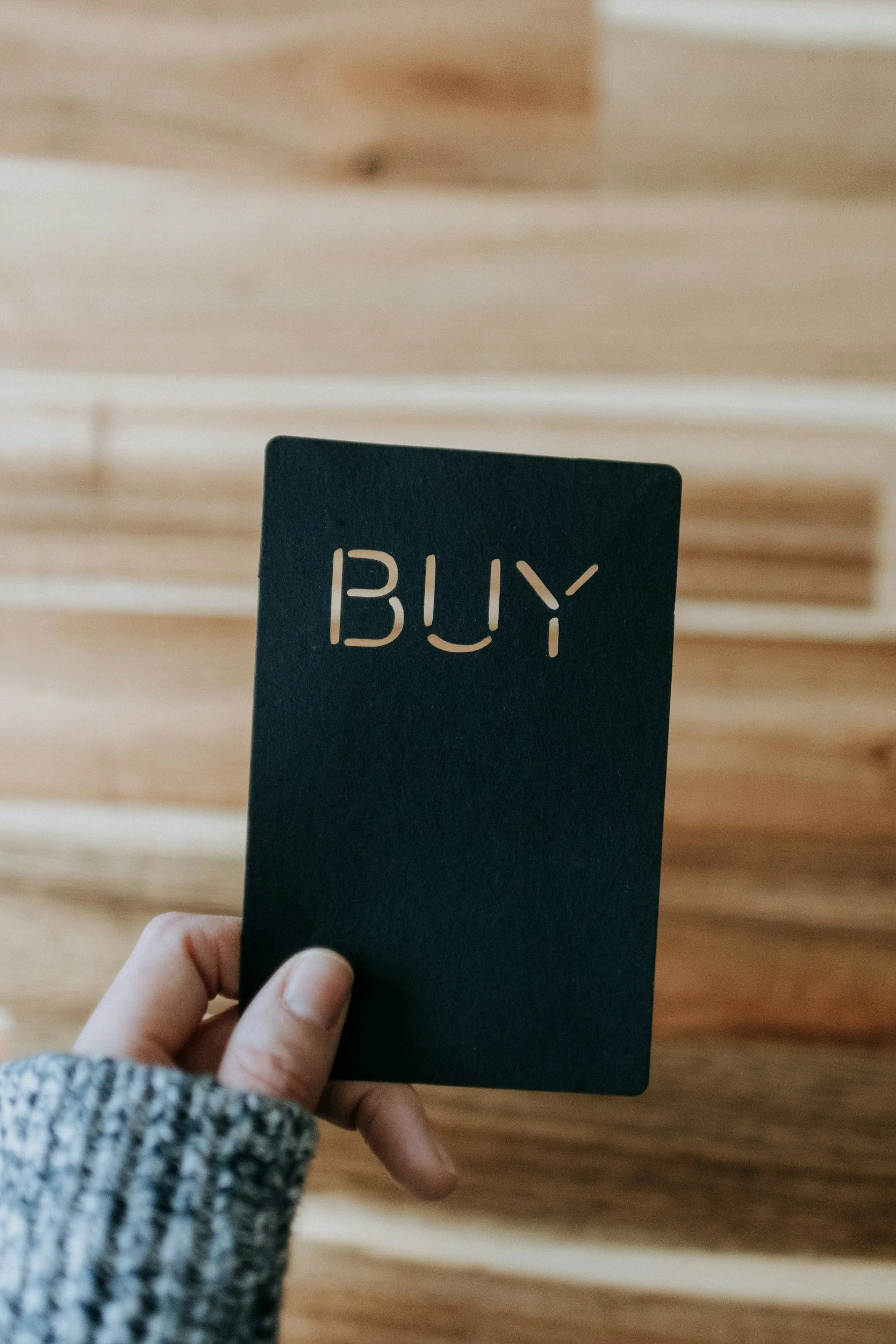 Person holding a black card with the word 'BUY' cut out, standing indoors with wooden flooring.