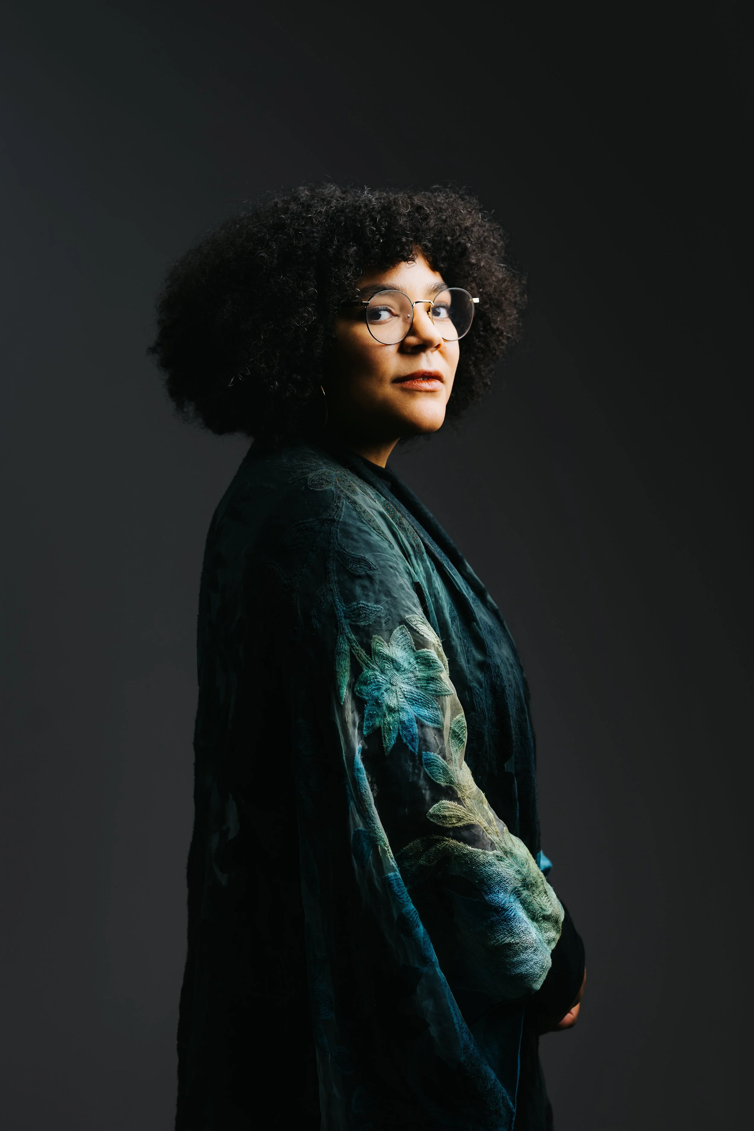 Portrait of a woman with curly black hair, glasses, and wearing a dark green floral jacket, against a black background.