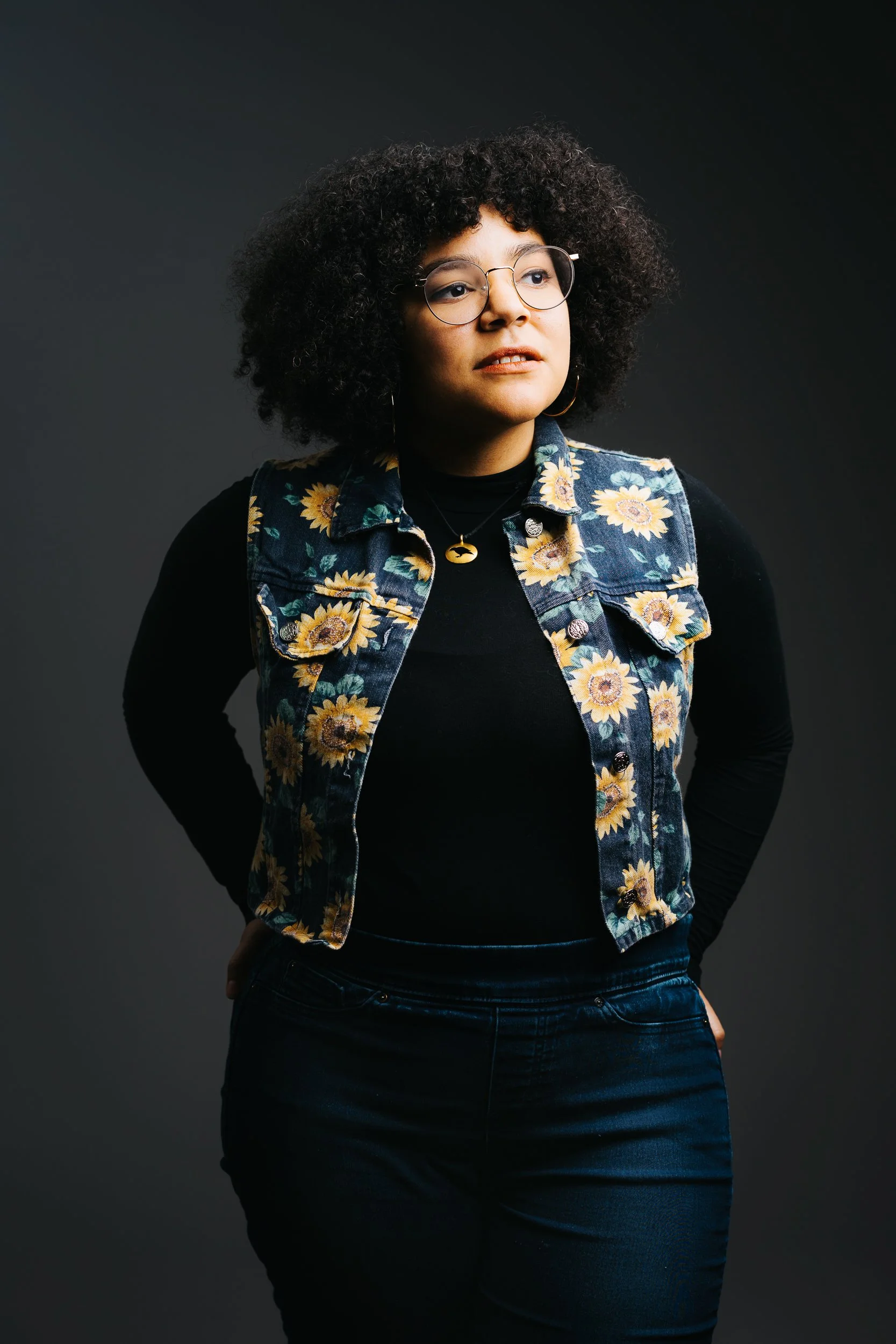 A woman with curly black hair wearing round glasses, a sunflower-patterned denim vest, and black clothing standing against a dark background.