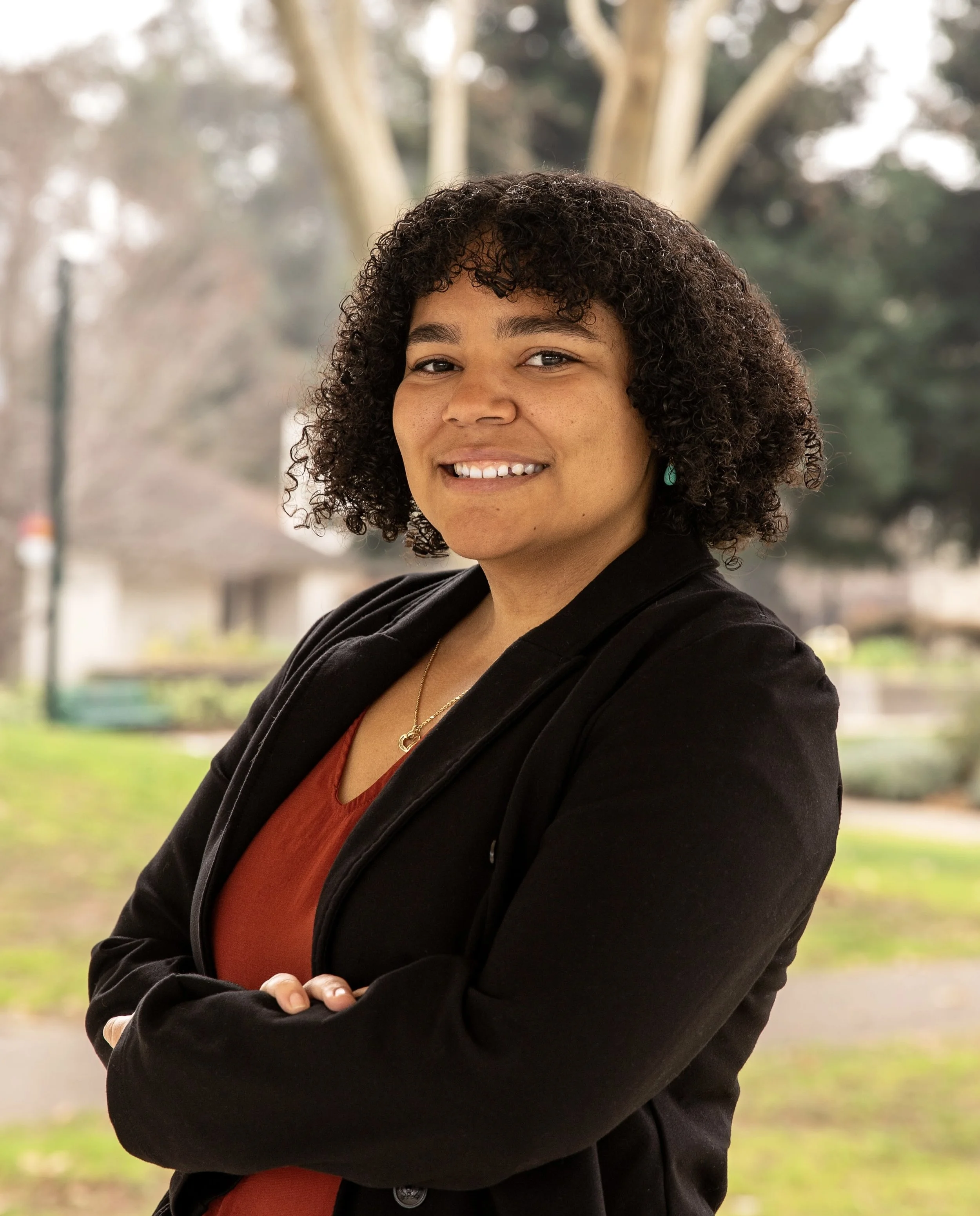 A woman with curly dark hair wearing a black blazer over a rust-colored top, smiling with her arms crossed outdoors in a park setting with trees and grass in the background.