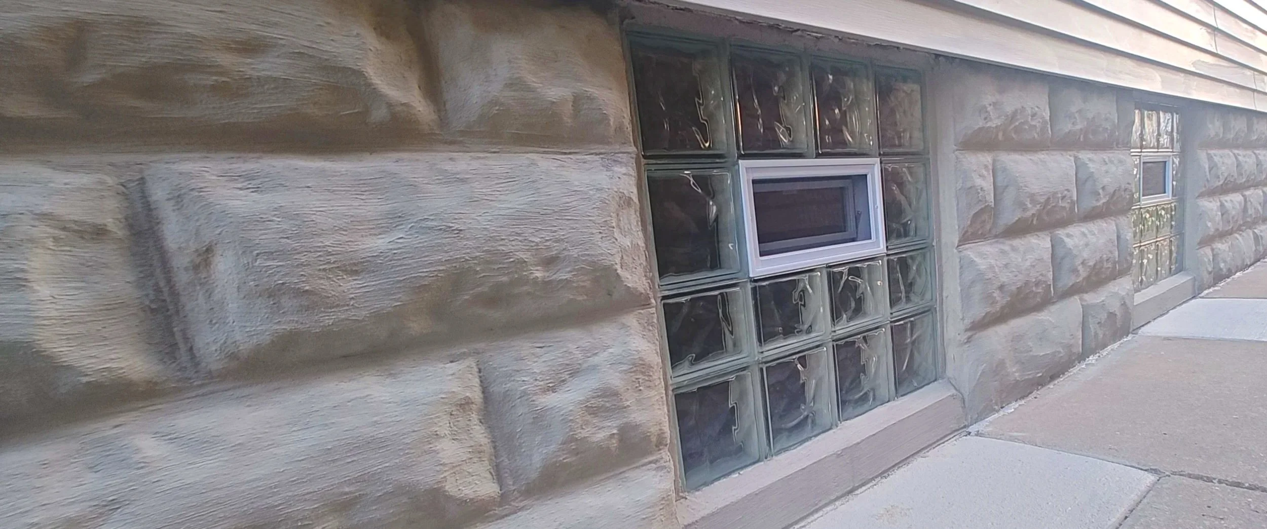 A section of a building's exterior wall made of rough-textured beige stone blocks with glass block windows and a small rectangular window, along with a concrete sidewalk.