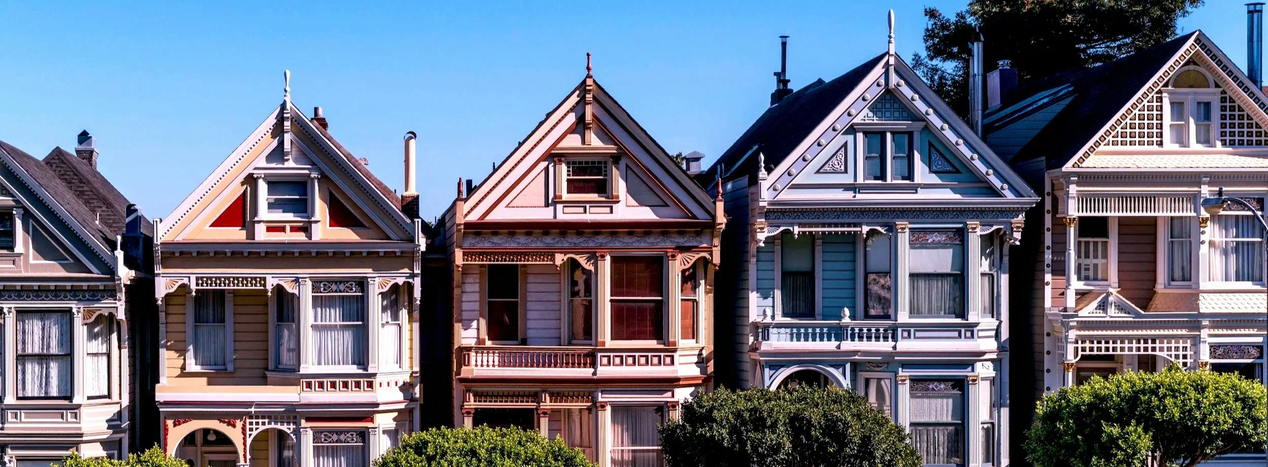 Row of Victorian-style houses with ornate woodwork, pointed roofs, and large windows under a clear blue sky.