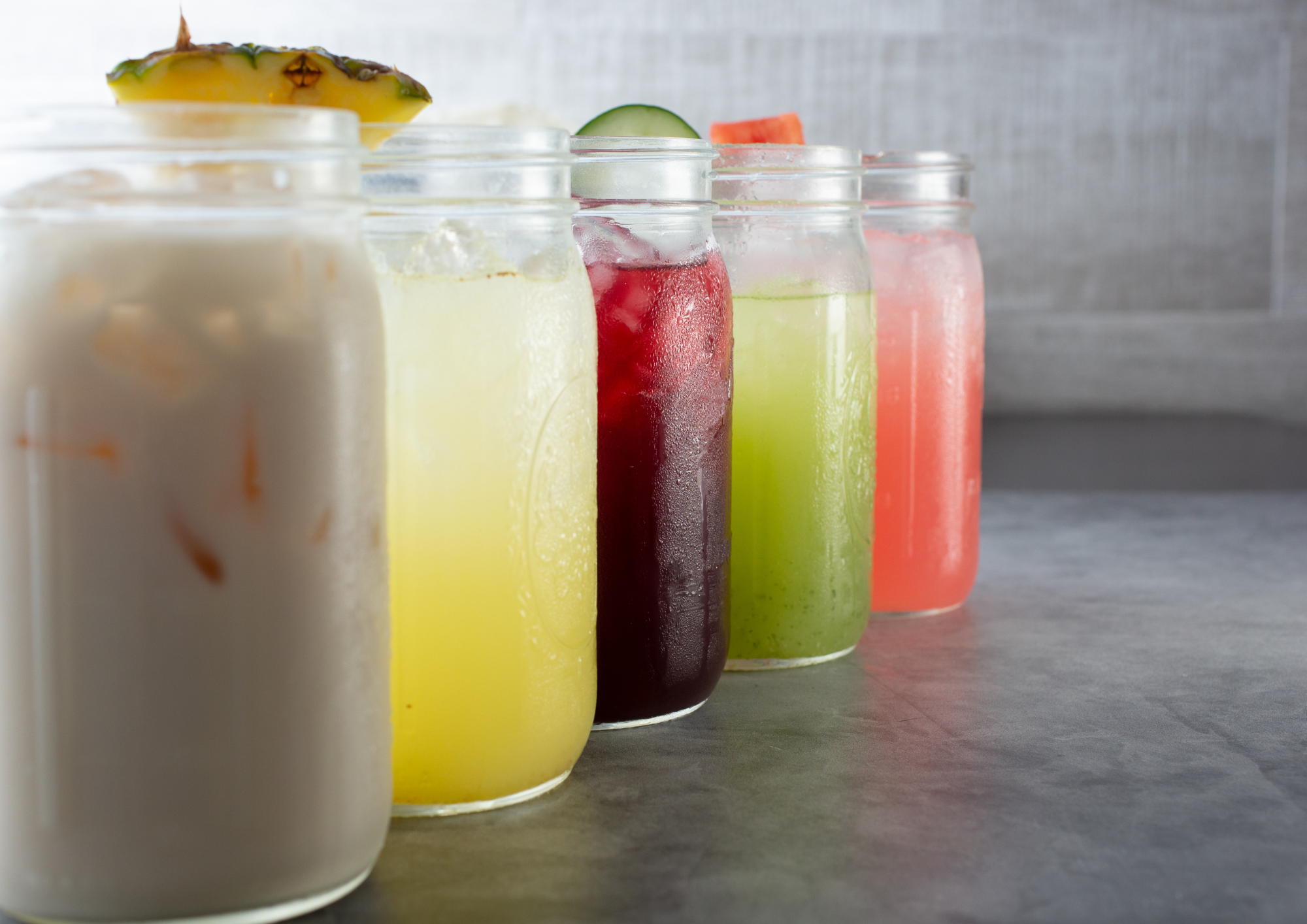 A row of colorful fruit smoothies in mason jars, with a pineapple on top of the first jar.
