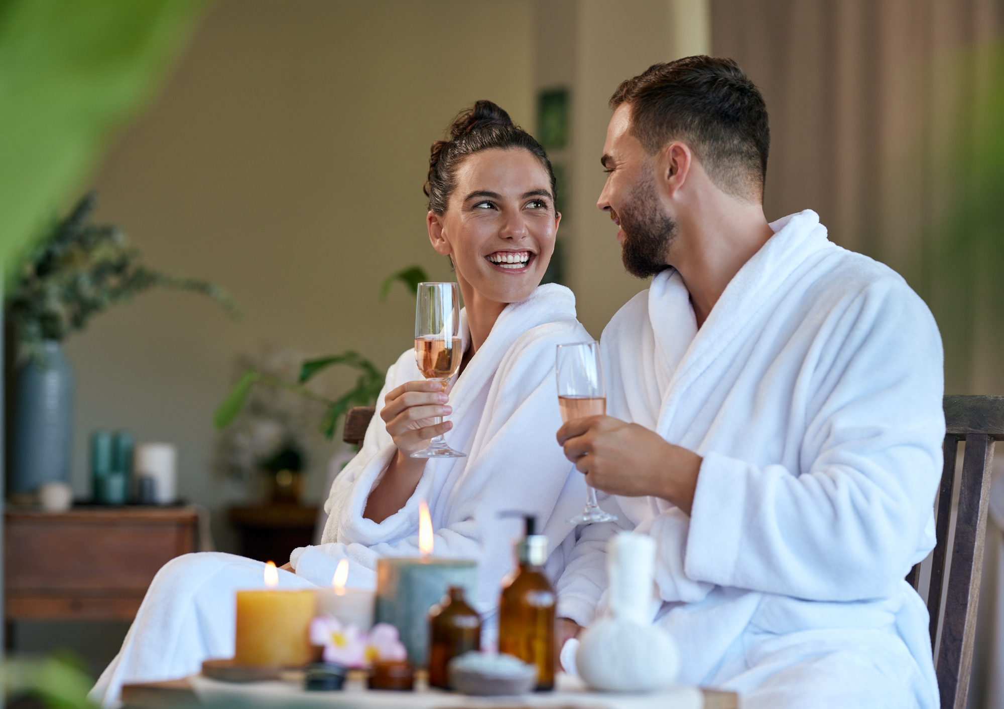 A man and woman in white bathrobes sitting close together, sharing a toast with glasses of rosé wine, smiling and looking at each other in a spa or wellness setting with candles and wellness products on a table in front of them.