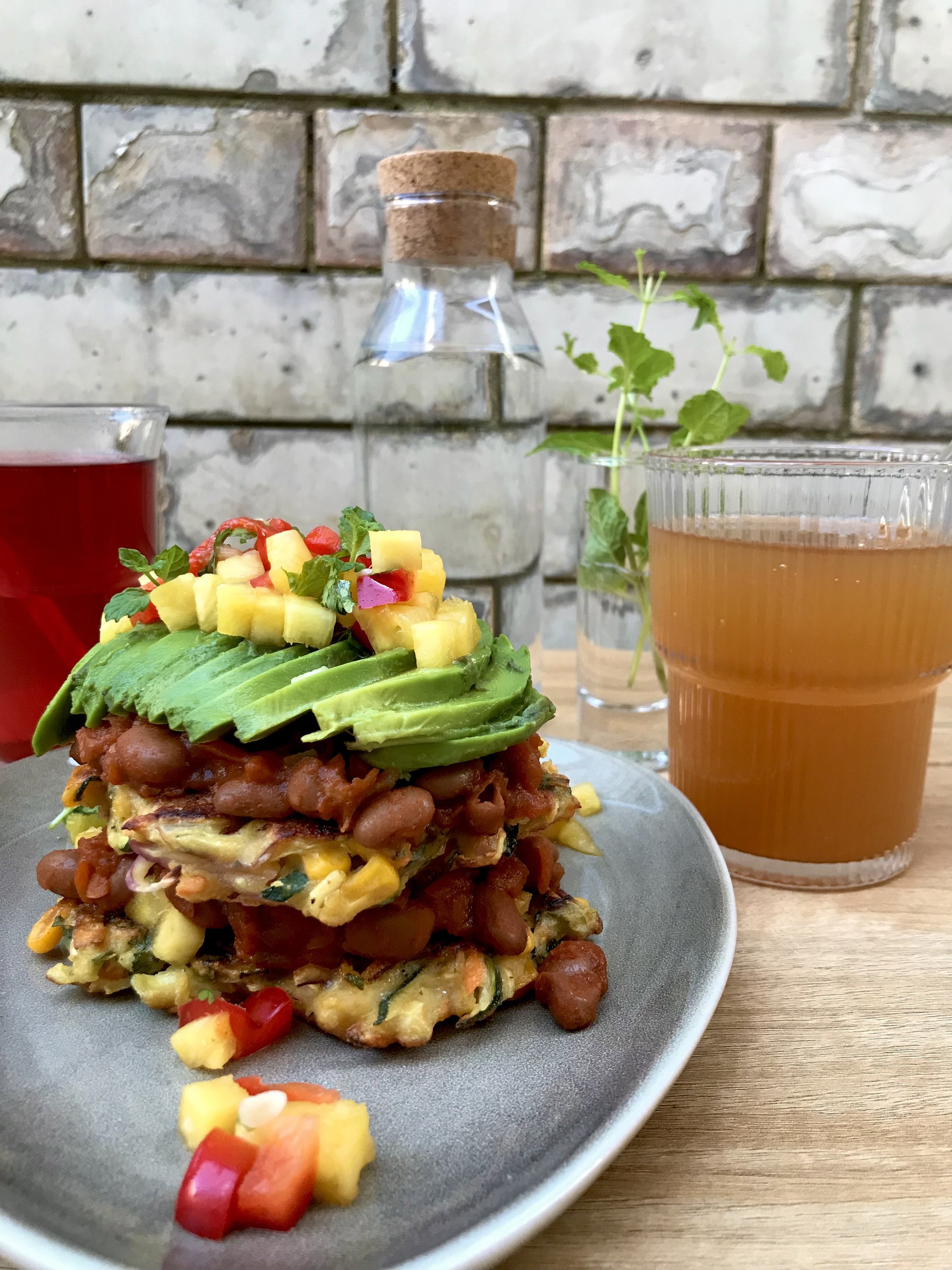 Layered vegetable pancake topped with sliced avocado, diced mango, and herbs, served on a gray plate with two glasses of red and pink beverages, a glass of water, and a small plant in the background.