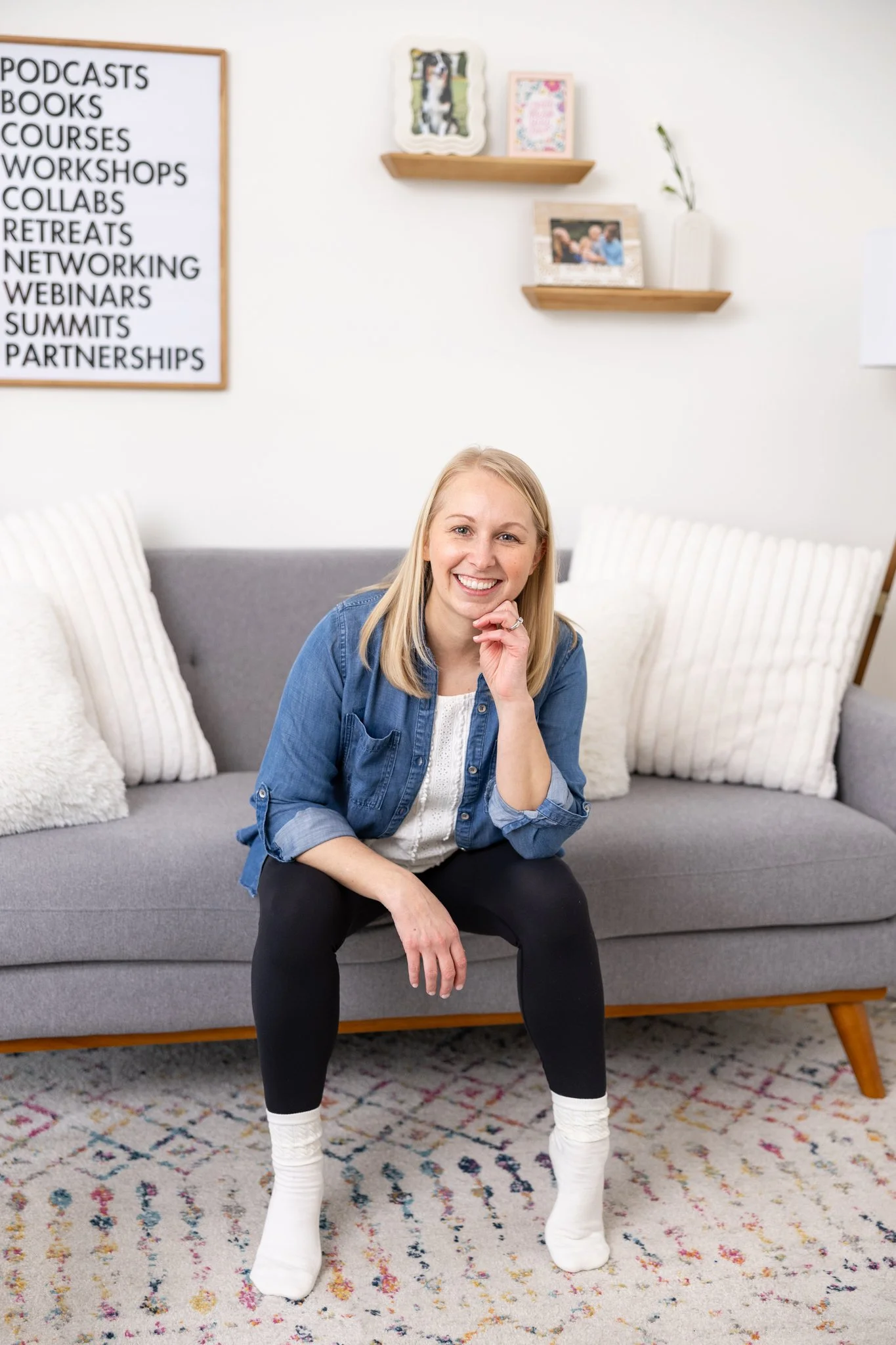 A woman sitting on a gray sofa in a living room, smiling, with white and cream pillows, a white wall with shelves, family pictures, and a sign listing various events and activities.