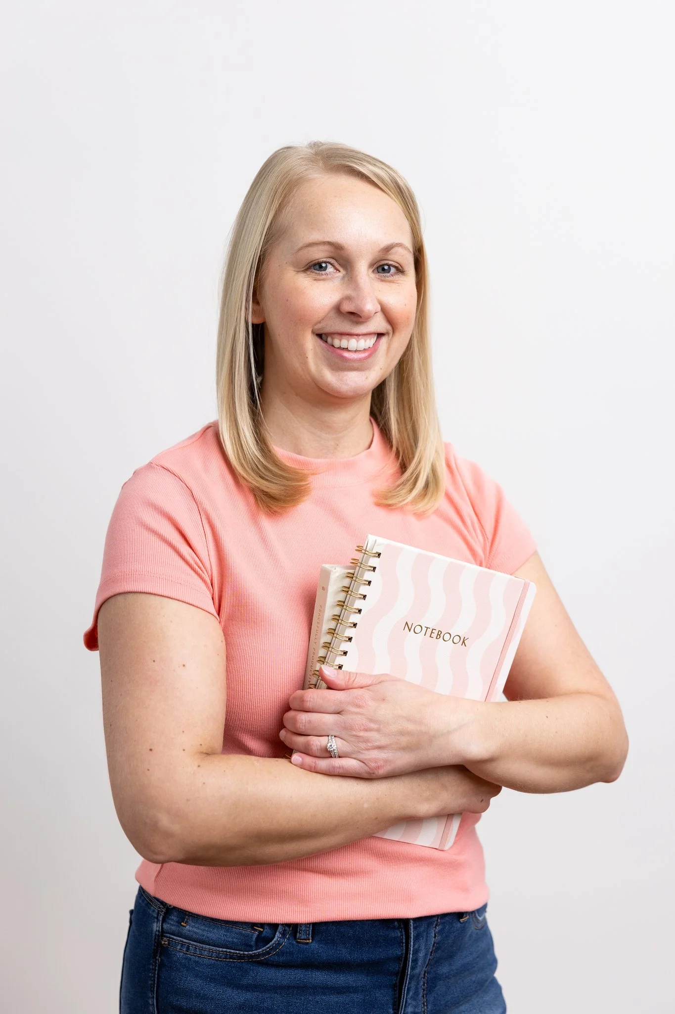 A woman with blonde hair, wearing a pink shirt and blue jeans, smiling while holding a pink notebook and a smaller notebook against a plain white background.