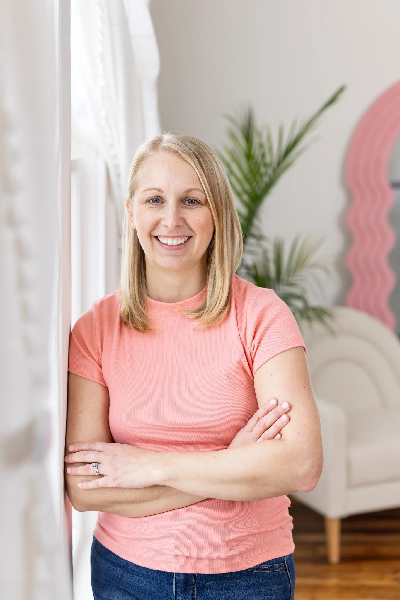 A woman with blonde hair, smiling, wearing a pink shirt, standing indoors with a green plant and pink decorative element in the background.