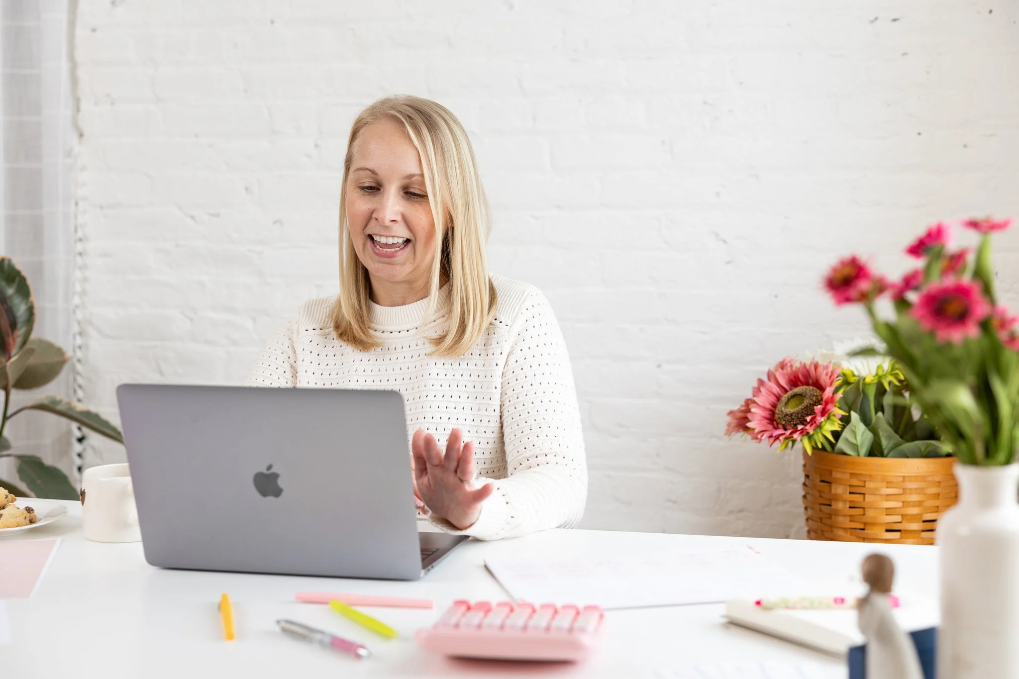 A blonde woman working on a laptop at a white desk, smiling and waving, with pink flowers in a basket in the background.