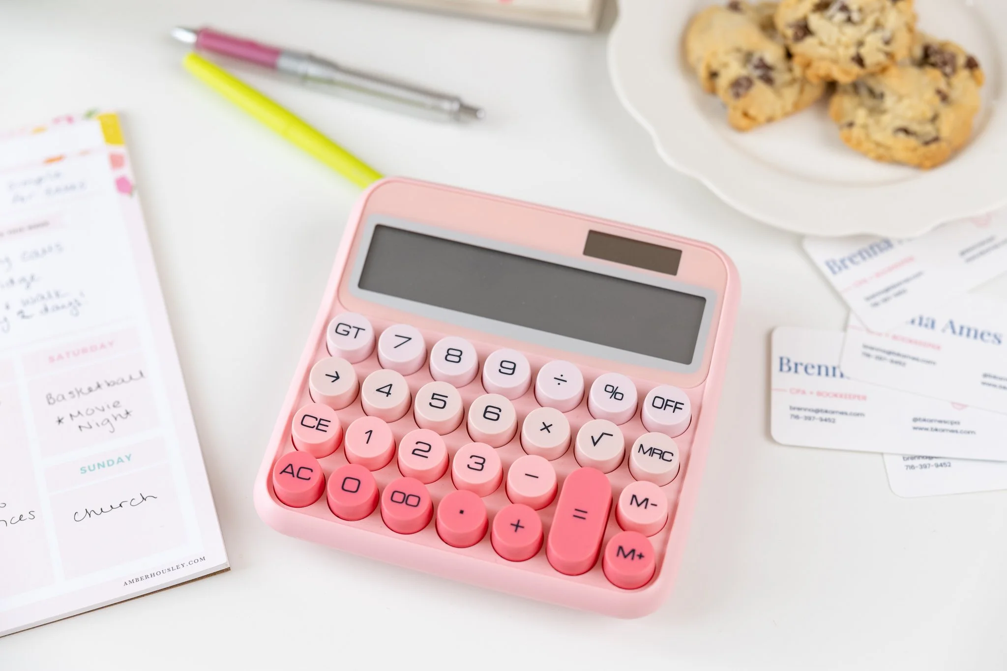 Pink calculator on a white desk with a notepad, pens, business cards, and cookies in the background.