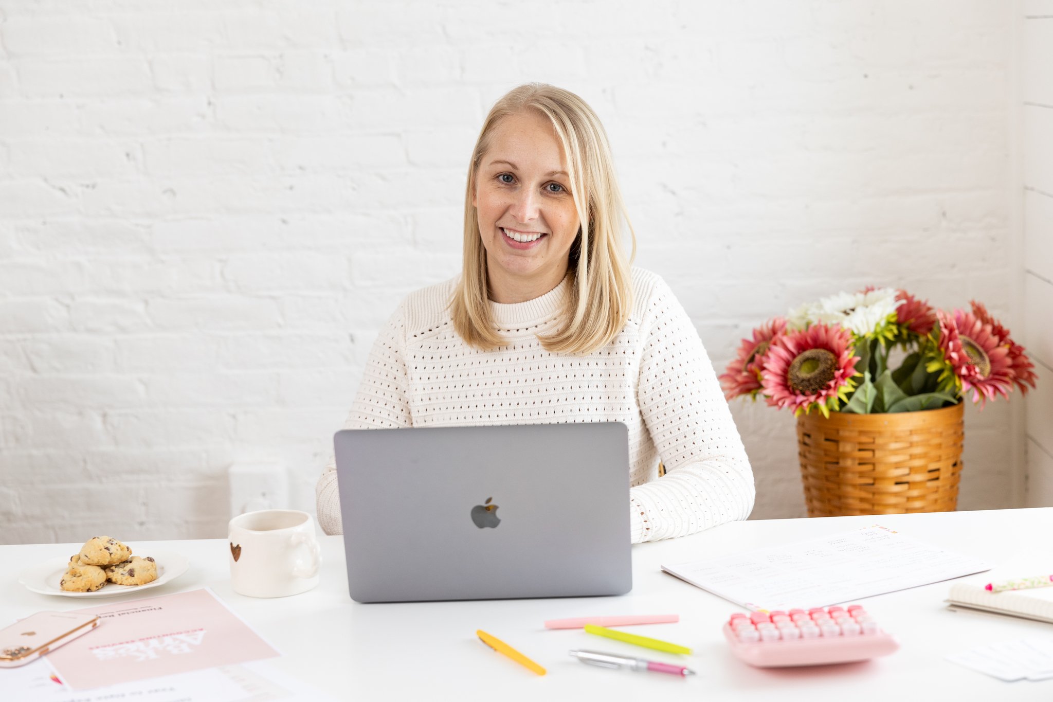 A woman with blonde hair sitting at a desk with a silver MacBook, a white coffee mug, a plate of cookies, colorful pens, a pink calculator, and a notebook with a pen, with a basket of flowers in the background.