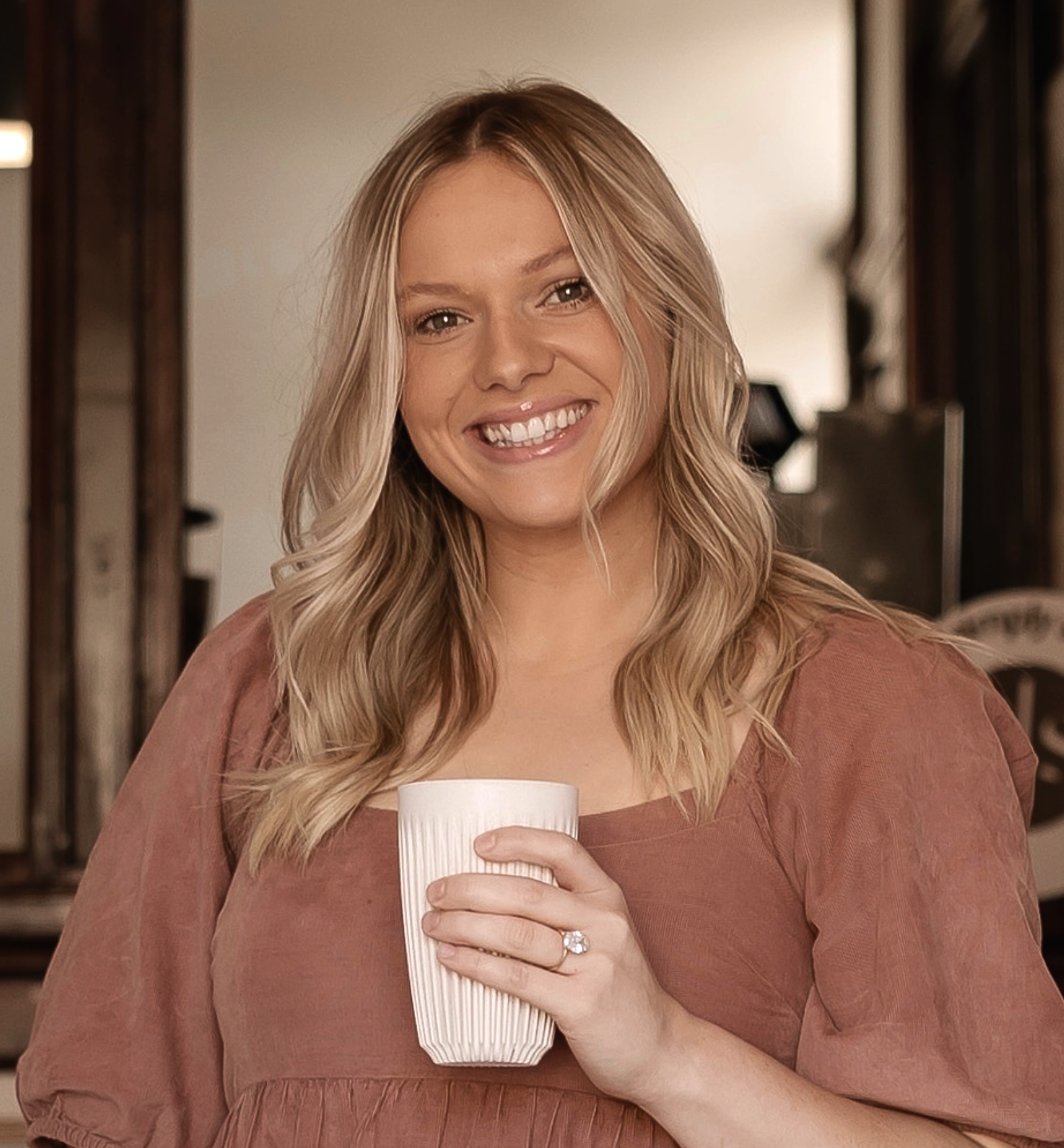 A young woman with wavy blonde hair smiling and holding a white striped coffee mug, wearing a rust-colored top, in what appears to be a cozy indoor setting.