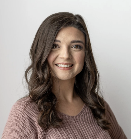 Smiling young woman with wavy brown hair wearing a pinkish sweater, against a plain white background.