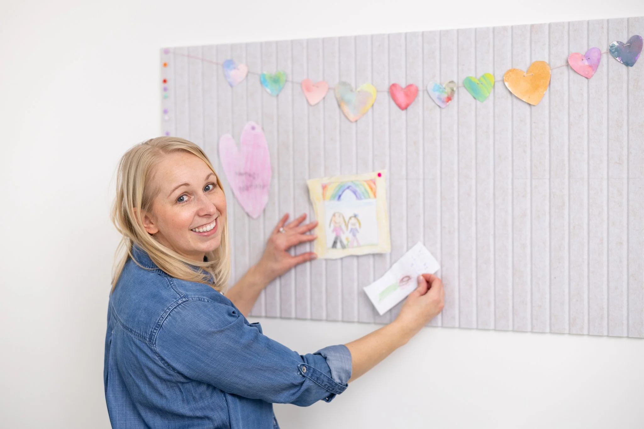 A smiling woman in a denim shirt pinning children's artwork to a gray fabric bulletin board decorated with colorful paper hearts.