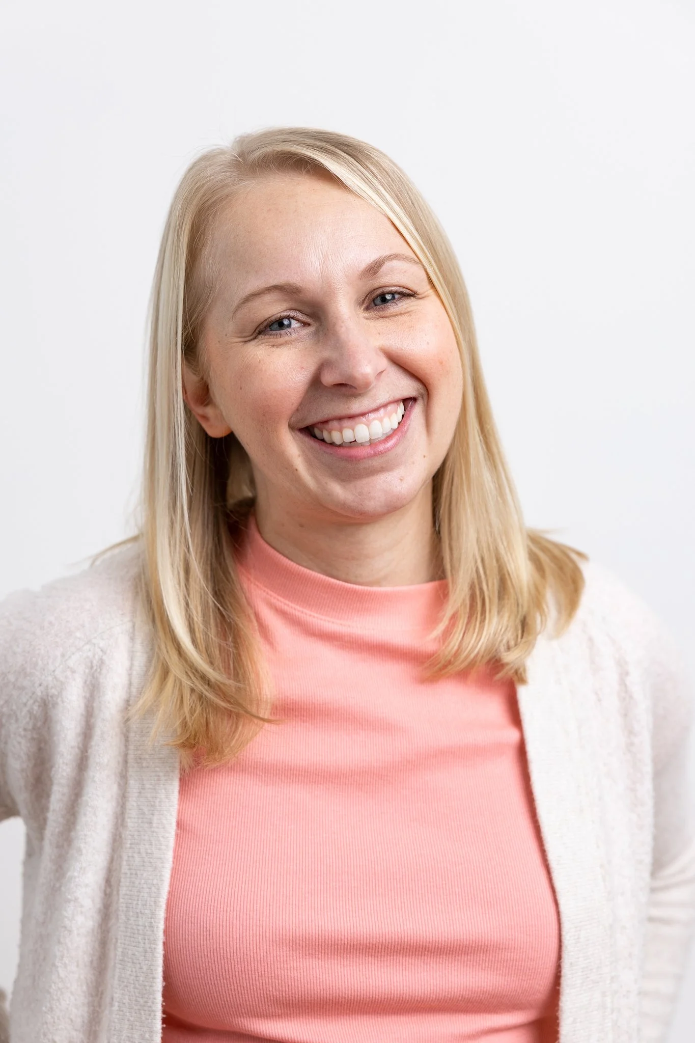 A smiling blonde woman with blue eyes wearing a peach-colored top and white cardigan against a plain white background.
