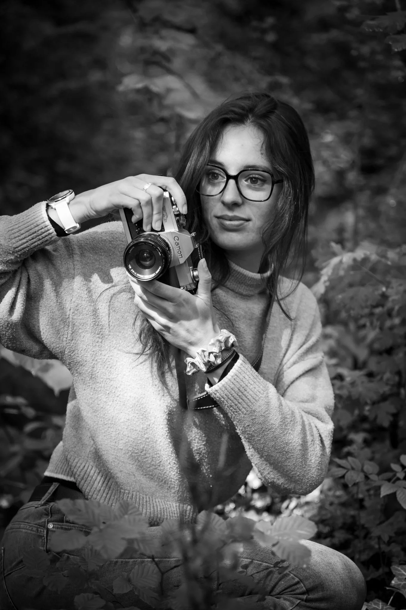 A young woman with glasses and long hair outdoors, holding a camera and looking to the side with a serious expression, surrounded by leaves and foliage.