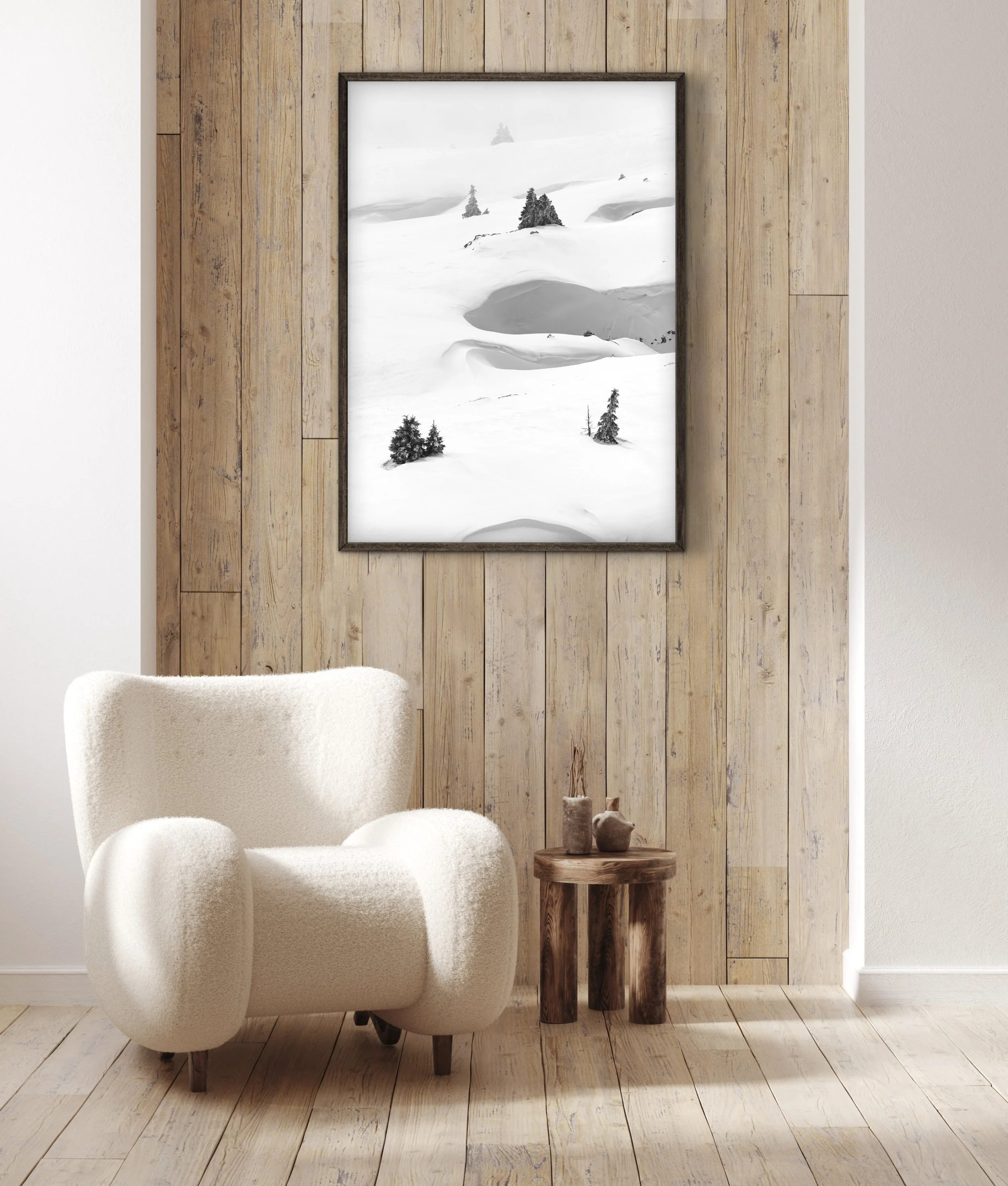 Interior of a room with a wooden accent wall, a white plush chair, a small wooden round table with two decorative vases, and a large framed black and white photograph of a snow-covered landscape with trees.