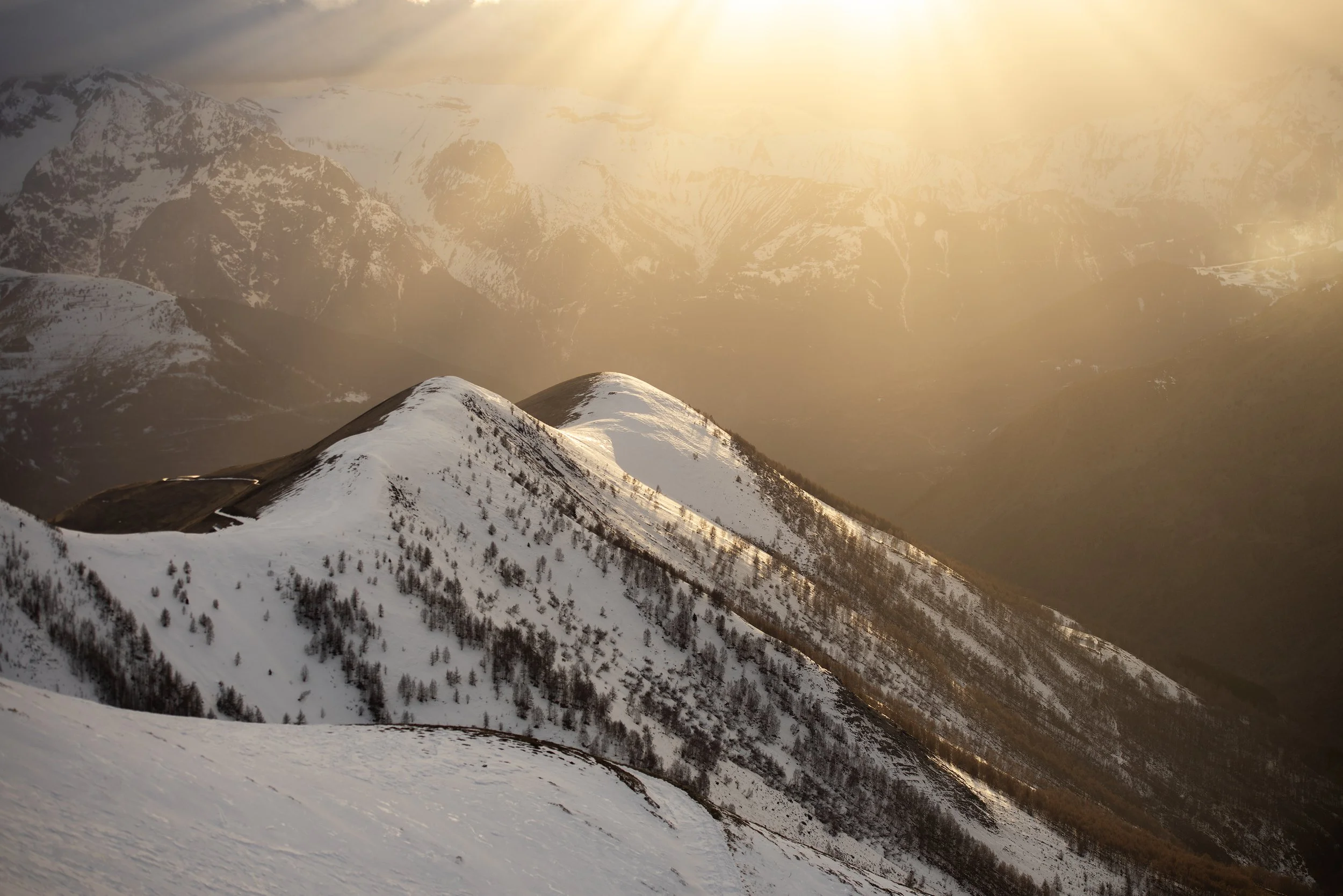 Snow-covered mountain range with cloudy sky and sunlight breaking through, creating a warm glow on the peaks.