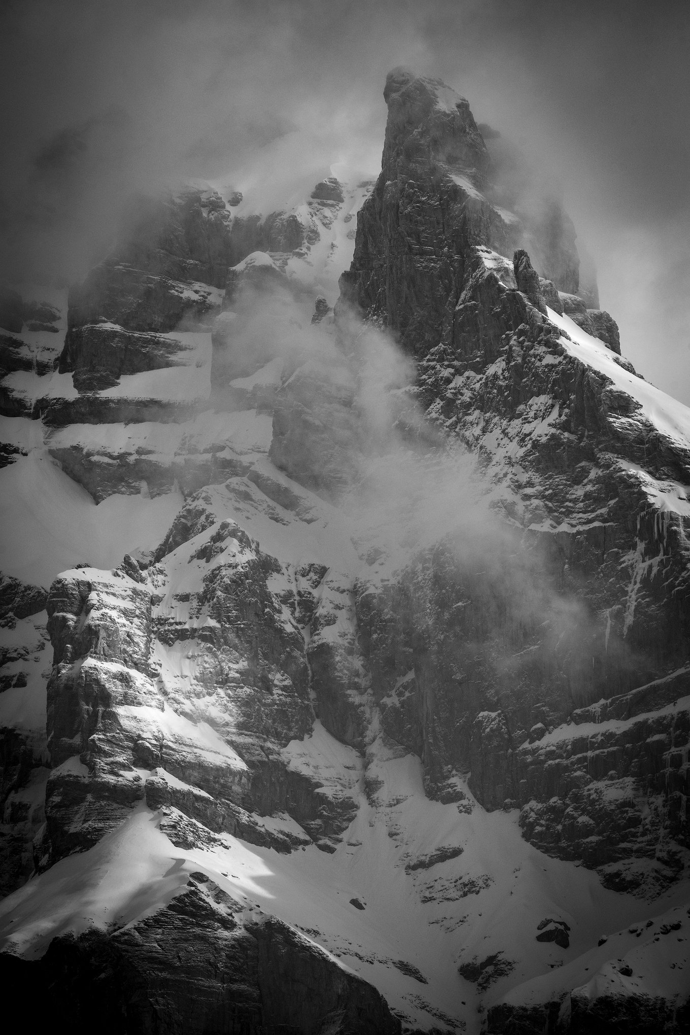 Snow-covered mountain peak shrouded in clouds and mist in black and white.