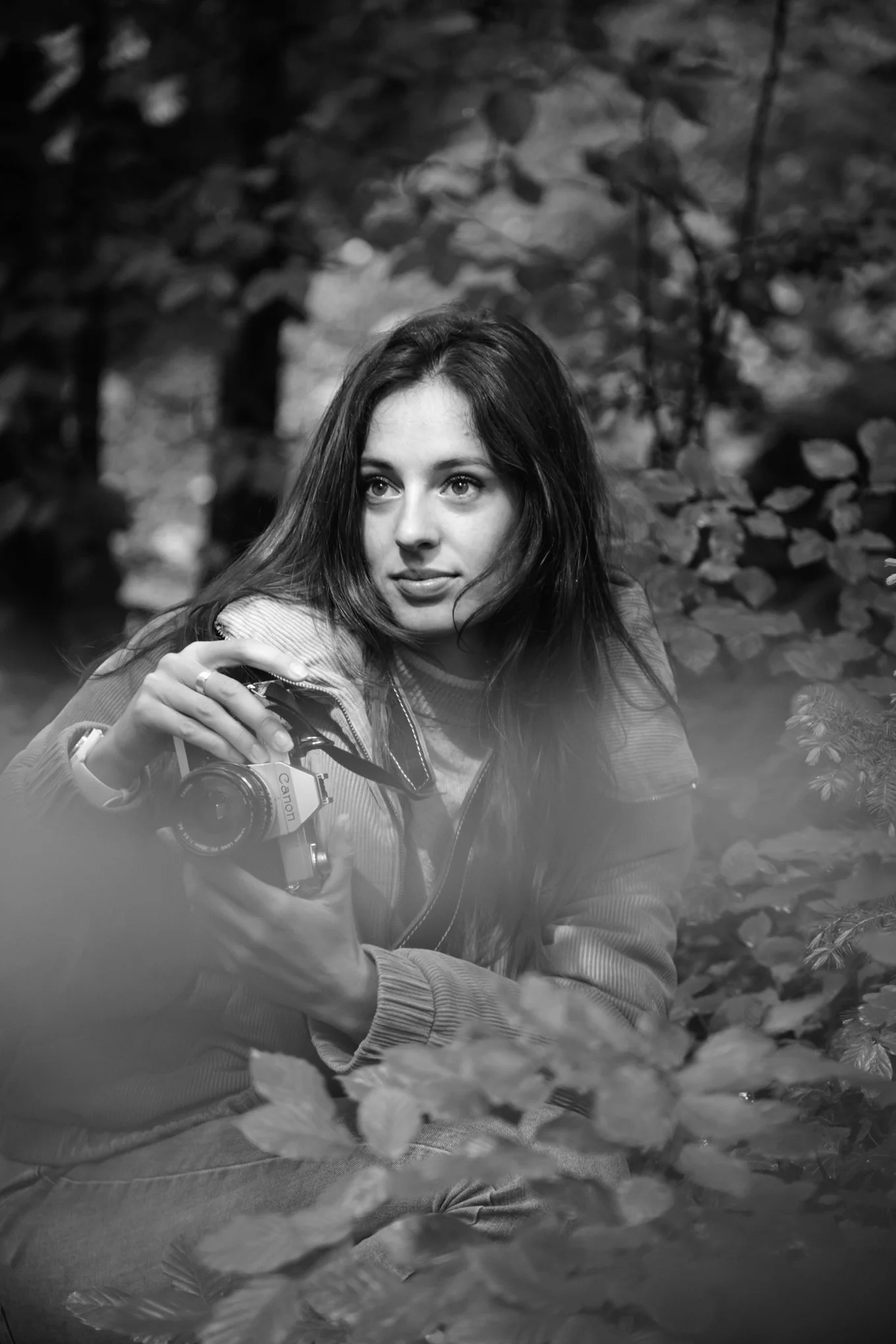 Black and white photo of a young woman with dark hair, sitting outdoors among foliage, holding a CANON camera, looking slightly off-camera.