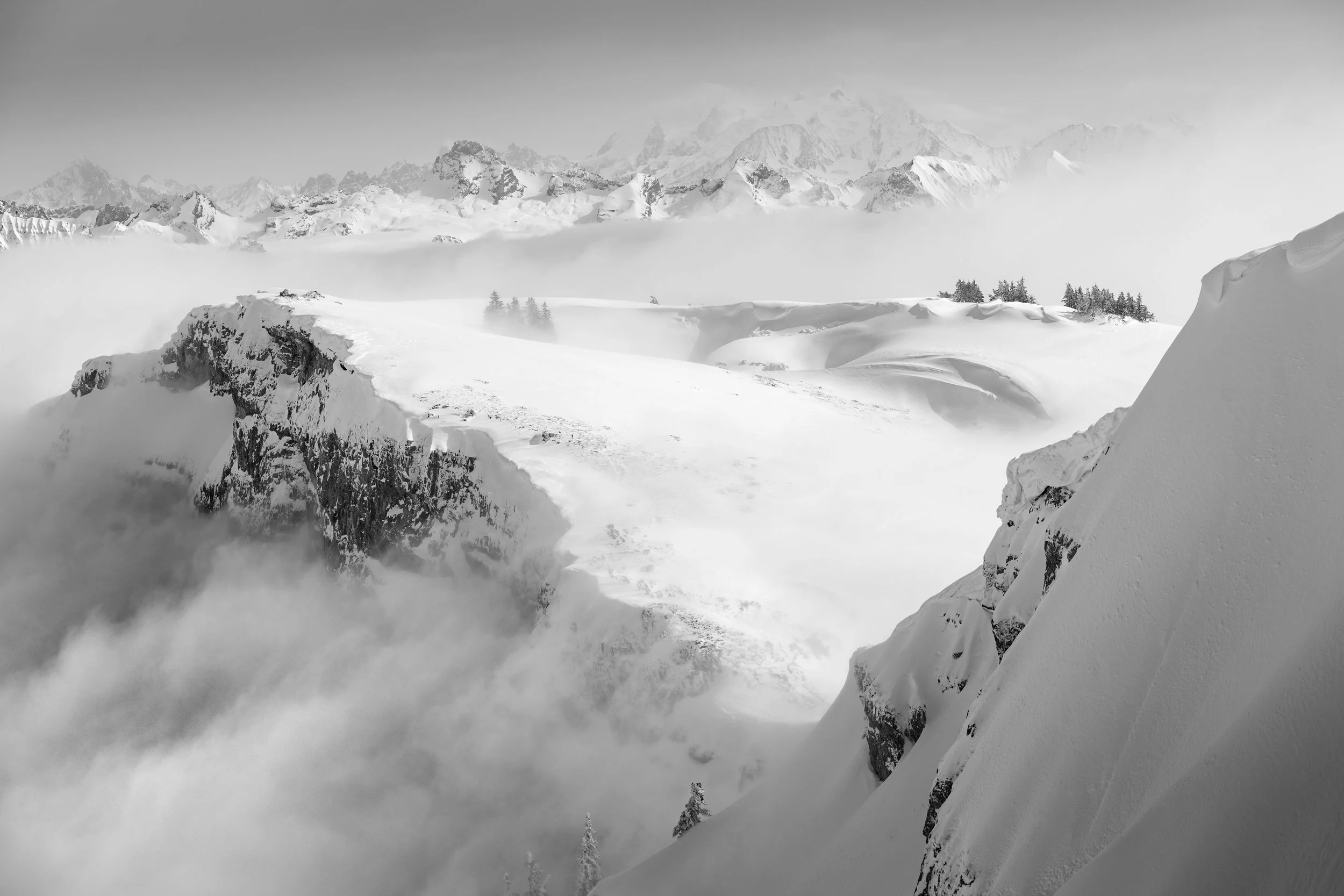 Snow-covered mountain landscape with peaks in the distance, fog or clouds in the valley, and snow drifts in the foreground.