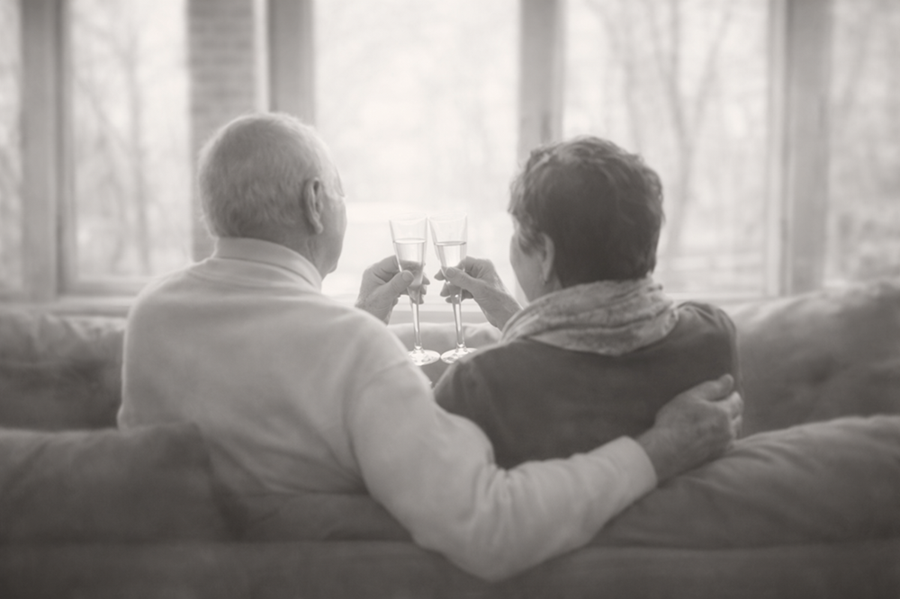 An elderly man and woman sitting on a couch, holding glasses of champagne, sharing a toast while facing a window with a view of trees.