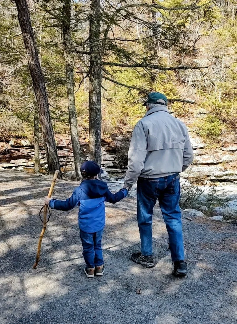 A man and a young boy walking hand in hand on a trail near a stream in a forest, with autumn foliage and tall trees around them.