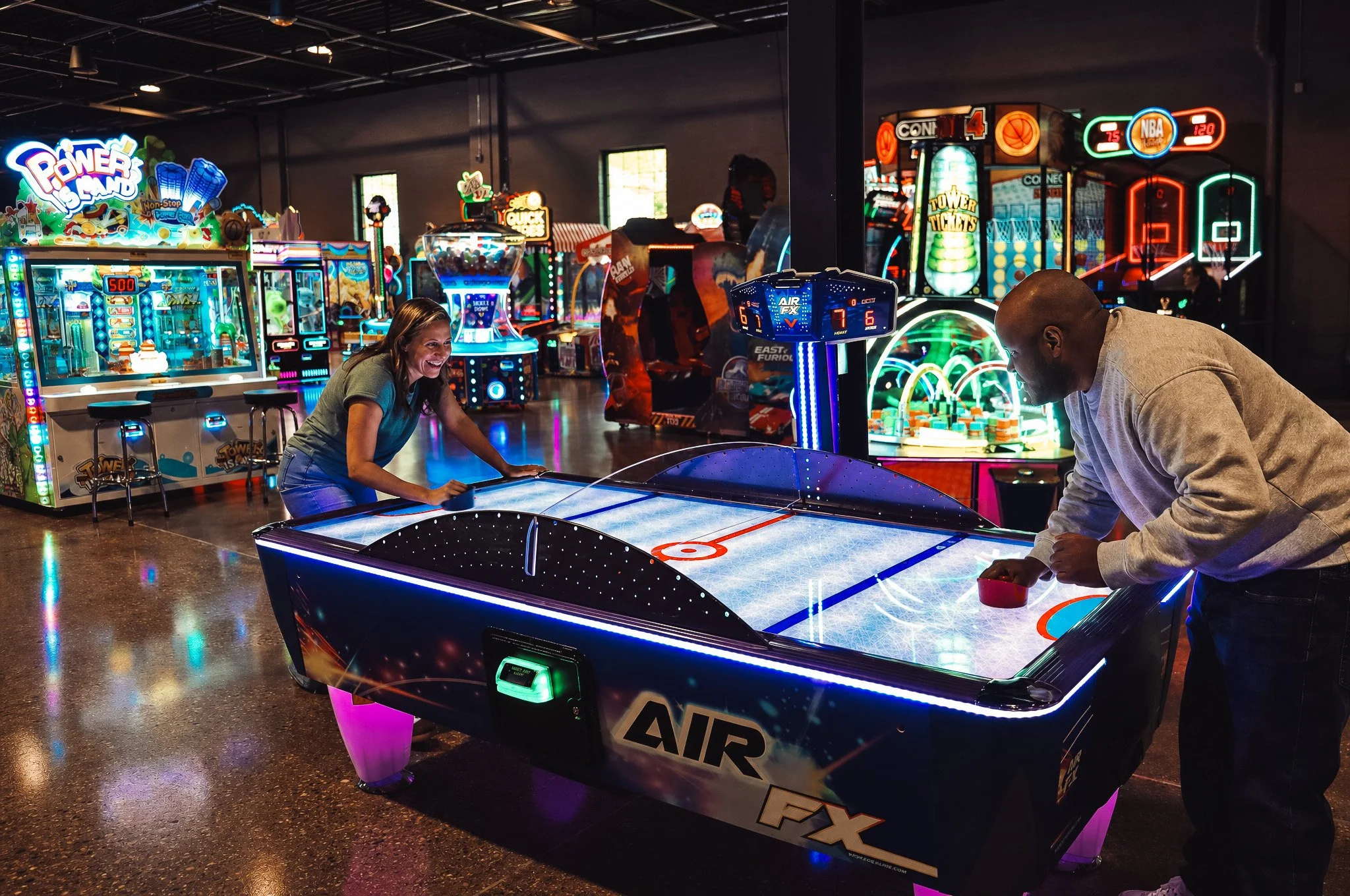 Two people playing air hockey at an arcade surrounded by colorful neon-lit arcade games and machines.