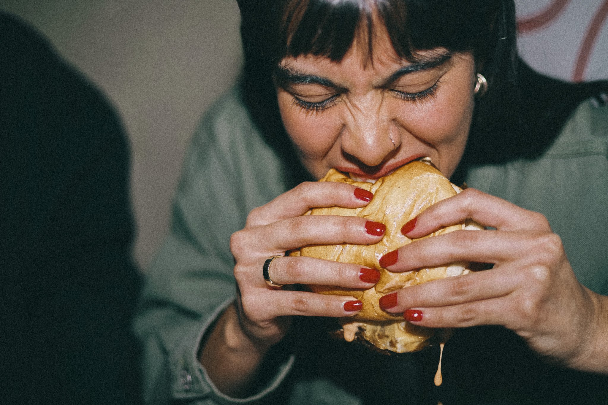 A woman with dark hair and red nail polish is biting into a sandwich with cheese, with some cheese visible dripping from the sandwich.