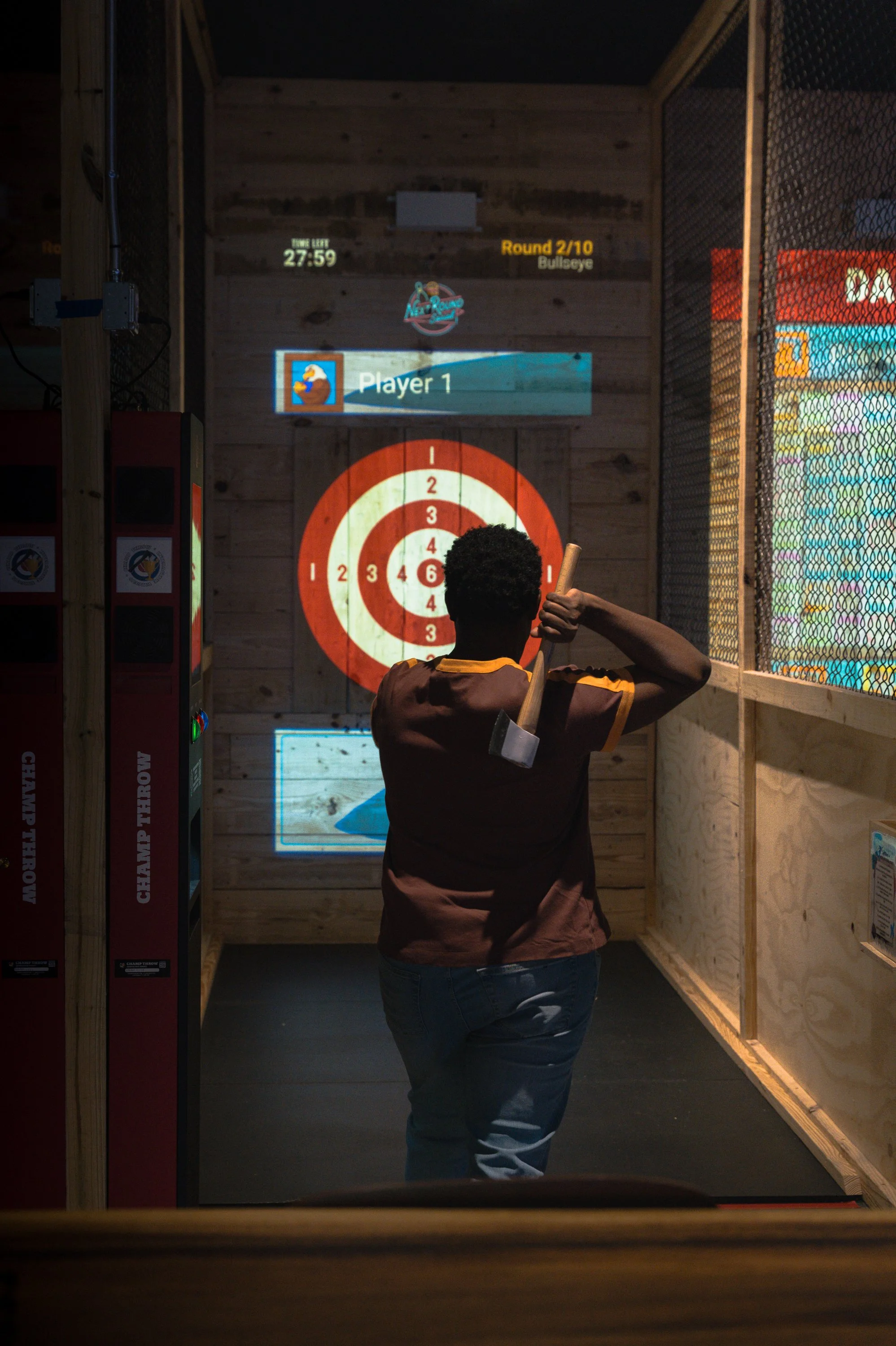 People playing axe throwing at an indoor venue, with target boards and digital score screens above.