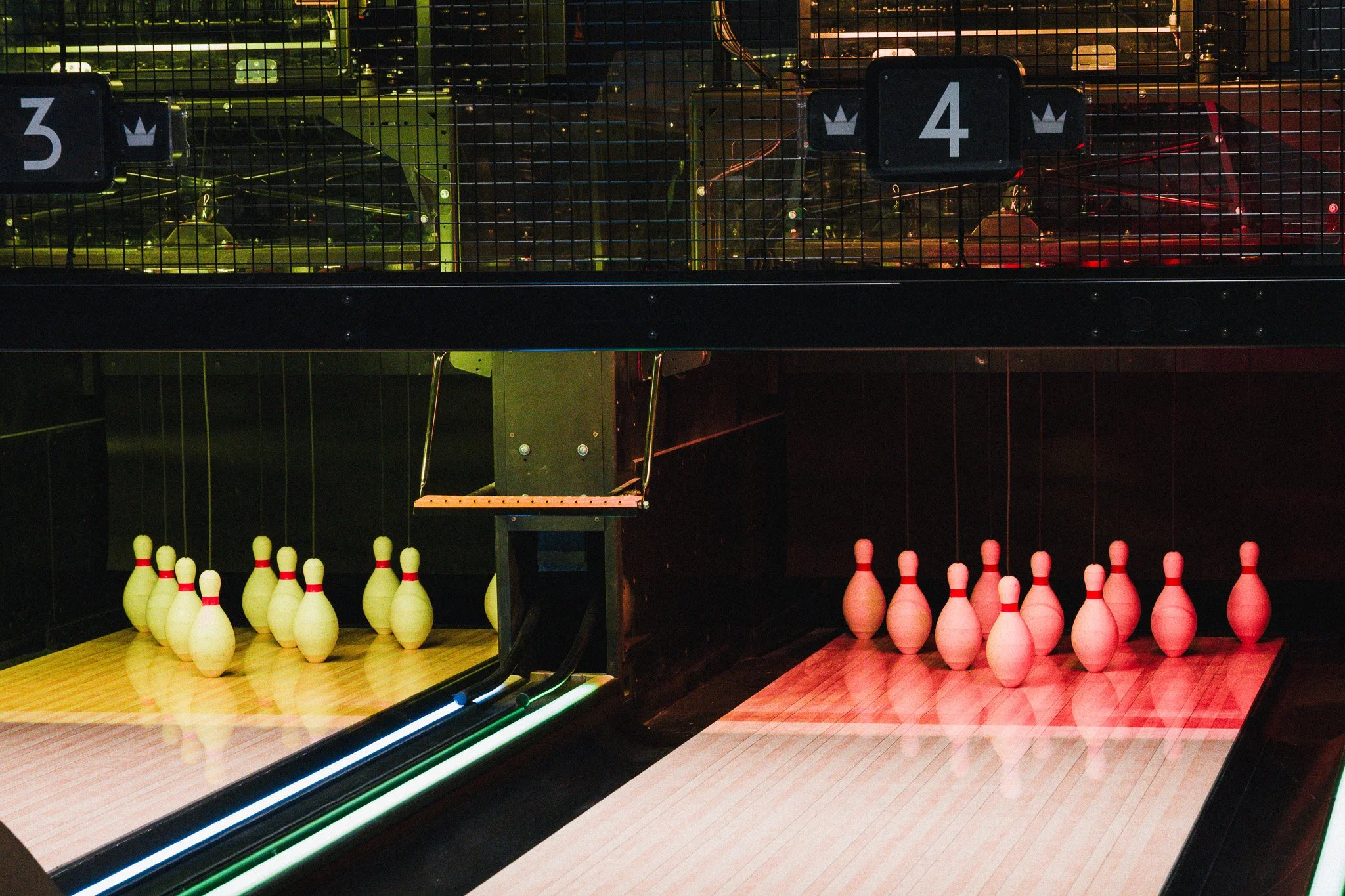 A lively indoor bowling alley with groups of people socializing and playing. The space features warm wood ceilings, modern lighting fixtures, framed landscape photographs with neon lighting on the dark wall, and seating areas with drinks.