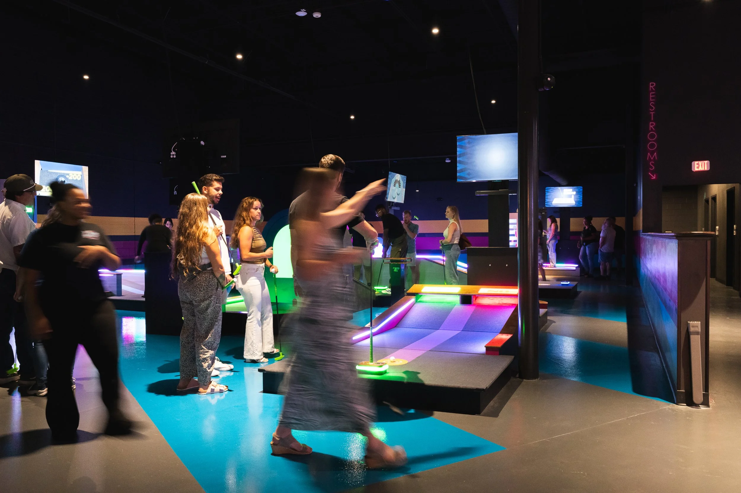 People playing miniature golf in a brightly lit, retro-themed indoor golf arcade with colorful neon lights and wall art of palm trees and sunset.