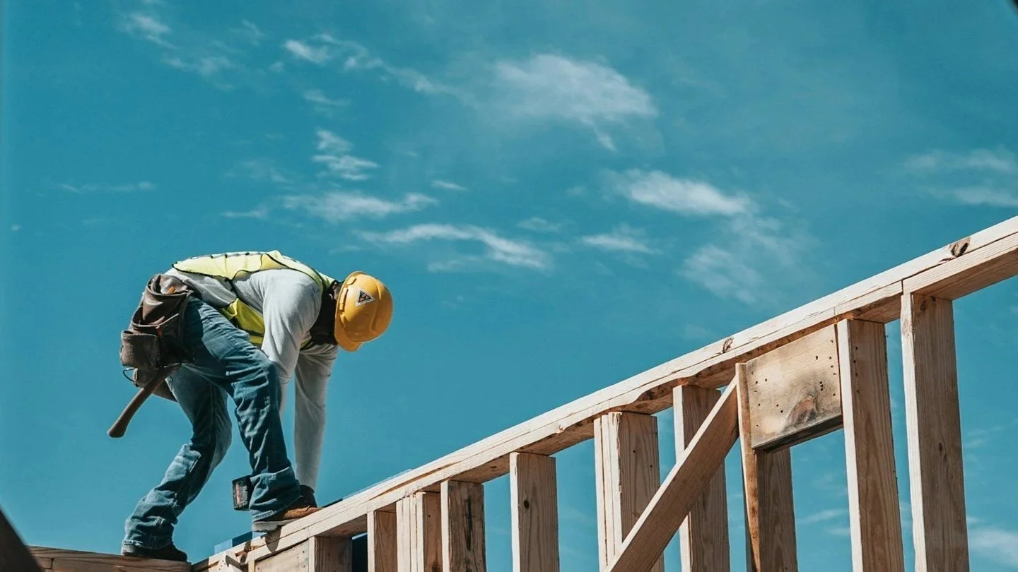 A construction worker wearing a yellow safety helmet and a reflective vest is working on a wooden framework for a building, with a blue sky and some clouds in the background.