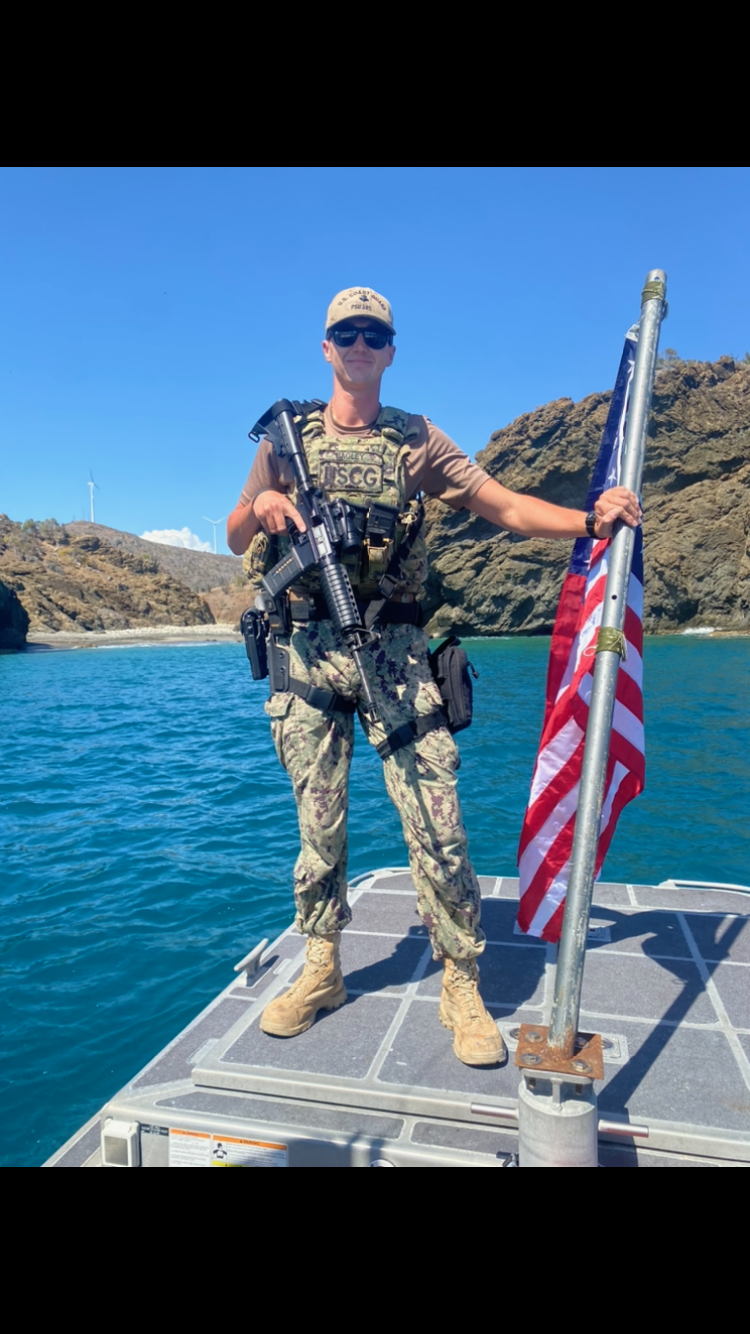A person in military uniform standing on a boat near rocky coastline, holding an American flag, with a firearm strapped to their chest, wearing sunglasses and a cap, under a clear blue sky.