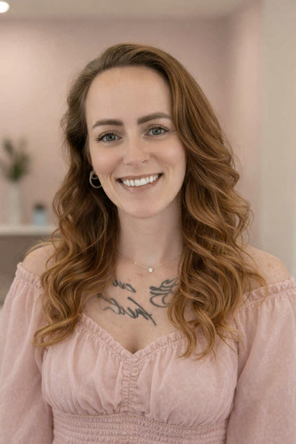 A woman with long, wavy red hair and blue eyes smiling, wearing a light pink off-shoulder dress, earrings, and a delicate necklace, indoors with a blurred background.
