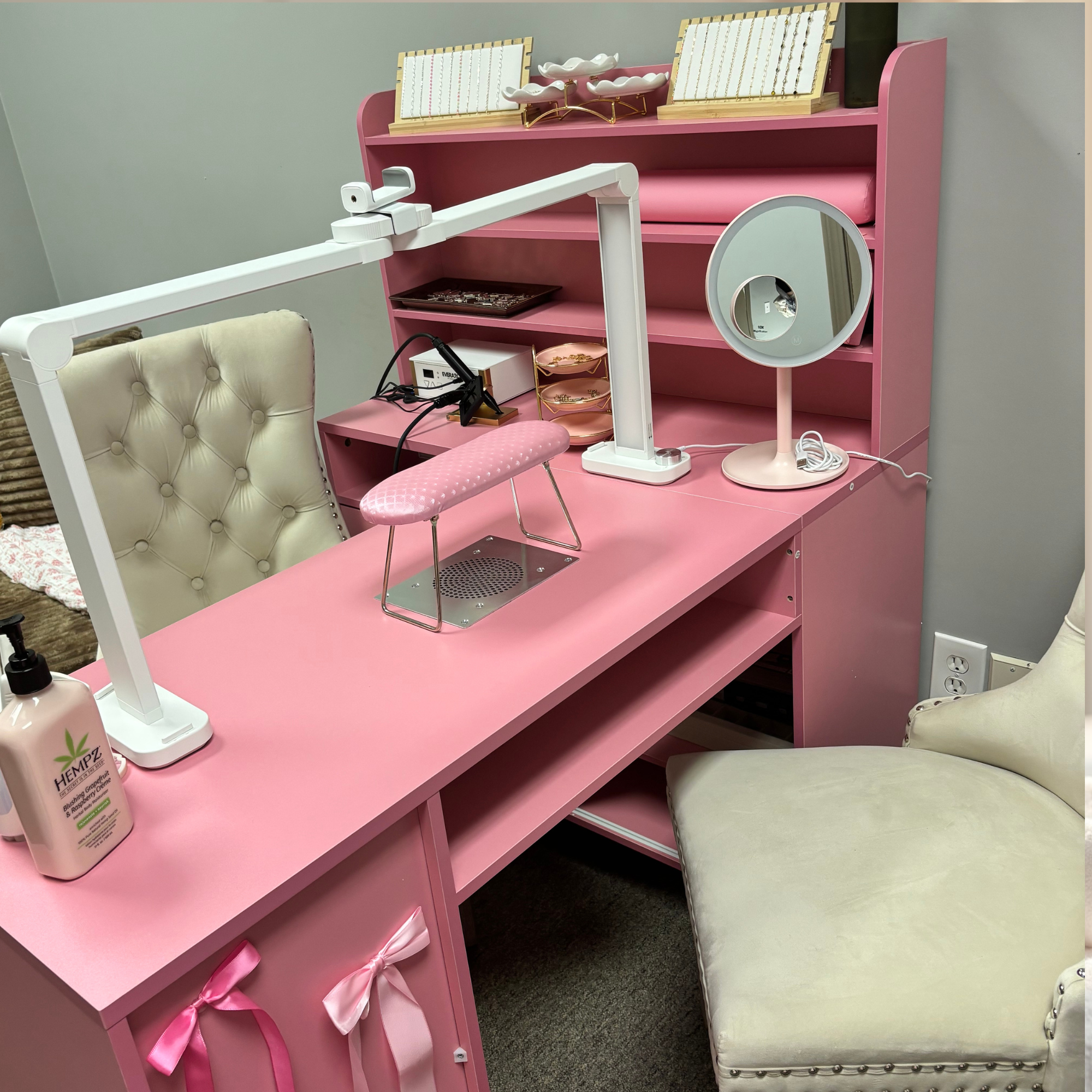 Pink manicure table with a pink cushioned nail filing mat, a lamp, a mirror, and a bottle of hemp lotion. There are white chairs with tufted backrests, and a pink shelving unit with various decorative items.
