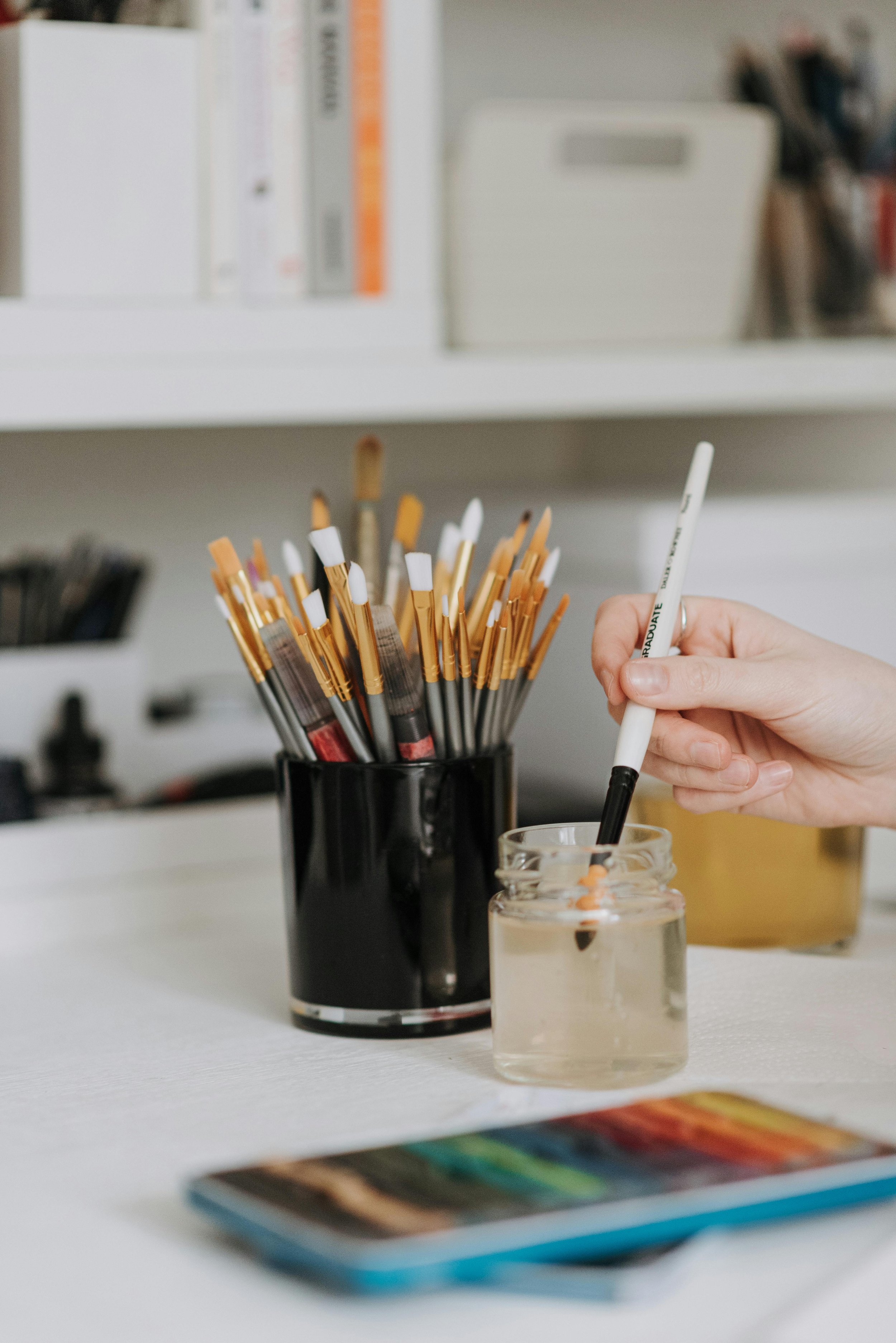A hand holding a black and white marker being dipped into a small glass jar of water, with a black cup filled with writing utensils in the background, on a white surface.