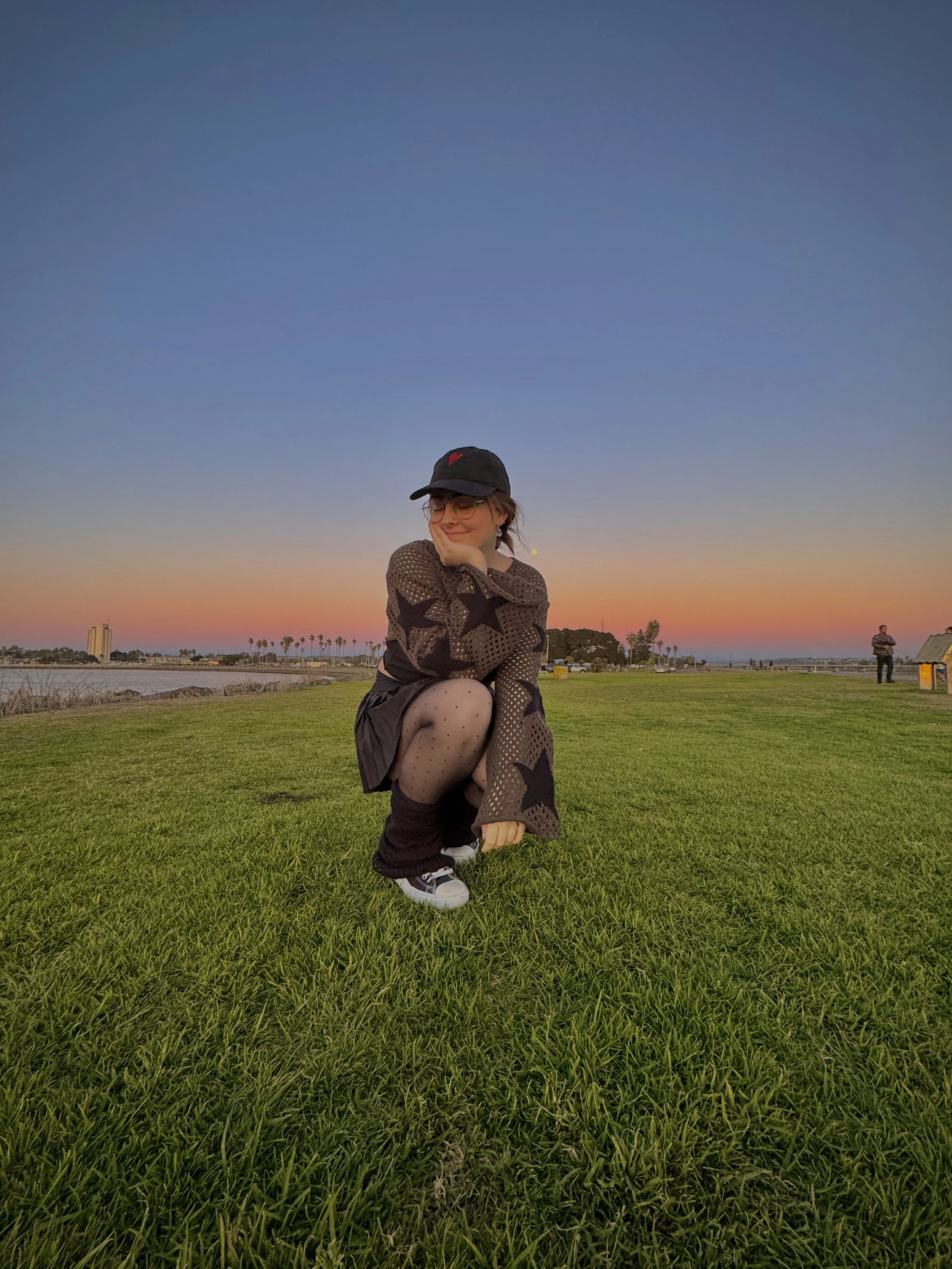 A woman crouching on a grassy field at sunset, wearing a black baseball cap, glasses, a patterned sweater with stars, black skirt, and polka dot tights.