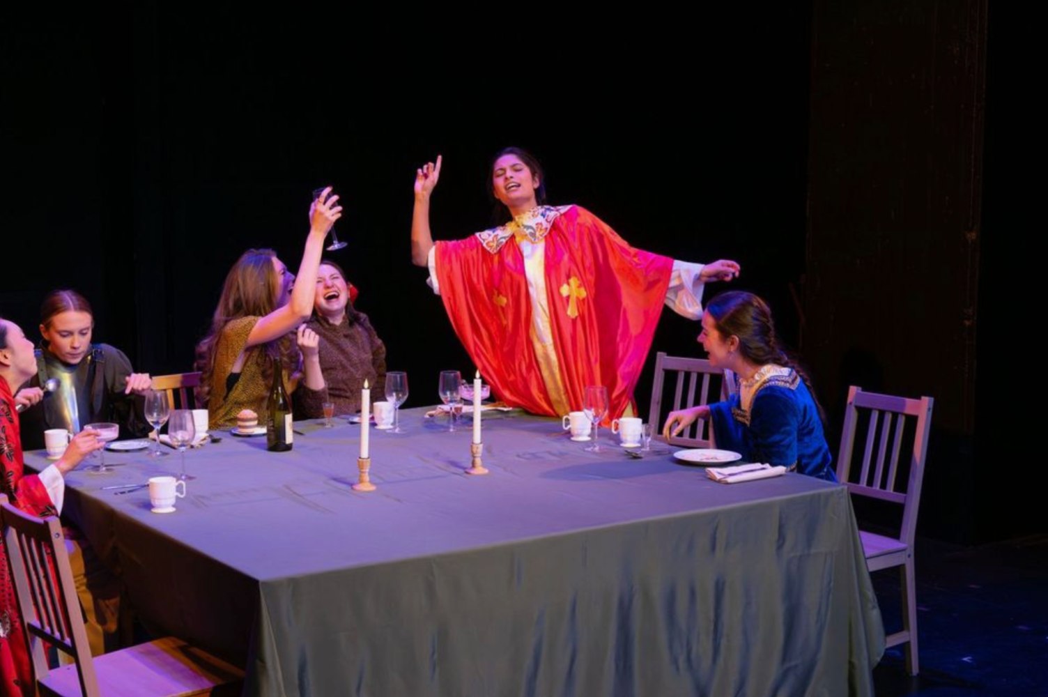 Women seated around a dining table at a celebration, with one woman standing and singing dramatically, dressed in a colorful cape, while others enjoy drinks and laugh in a festive setting.
