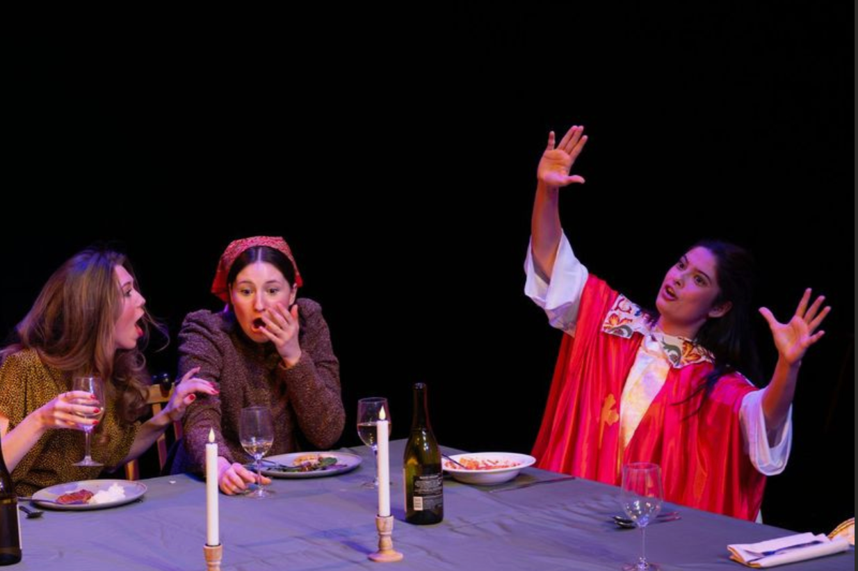 Three women at a dinner table, two of them are reacting with surprise or shock to the third woman, who is dressed in colorful, theatrical clothing and appears to be performing or speaking dramatically.