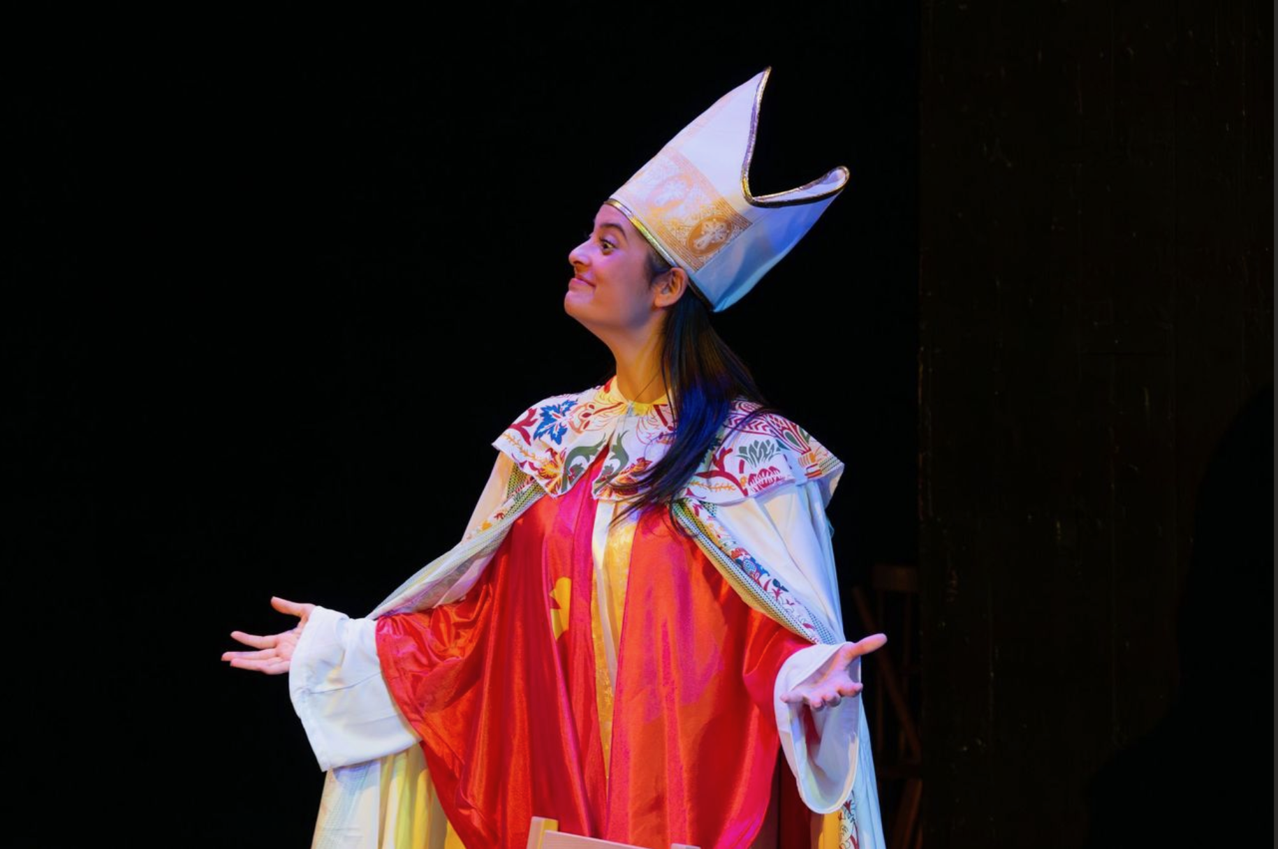 A woman on stage dressed in traditional costumes, wearing a tall white headdress with gold accents, and extending her arms outward with a smile.