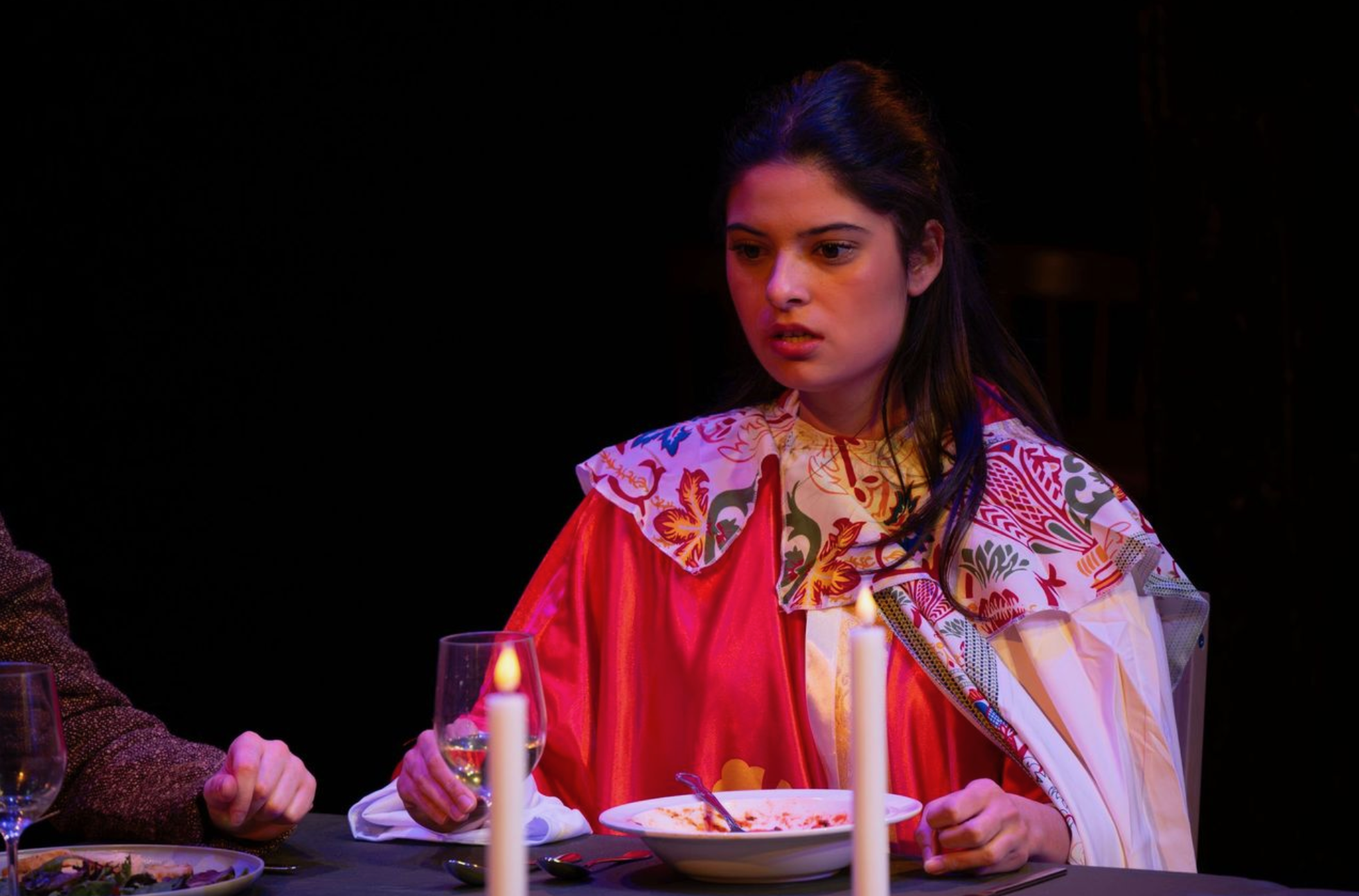 A young woman with dark hair sits at a dinner table with a serious or contemplative expression, wearing a colorful, patterned bib and a red apron. The scene is dimly lit with candles, and she has a plate of food in front of her.