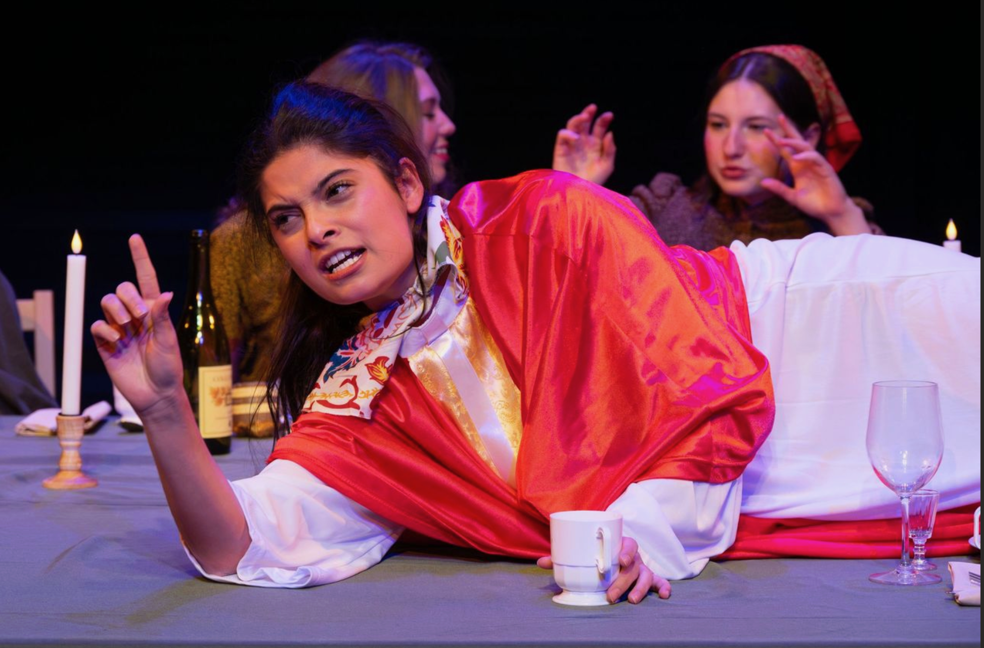 Three women sitting at a table during a theatrical performance. The woman in front is lying on her side, wearing a red satin robe and making a facial expression of frustration or annoyance, with one finger raised. Behind her, two women are in the background, one smiling and the other with her hand near her face, both listening intently. There are lit candles, a wine bottle, glasses, and cups on the table, indicating a dinner scene.