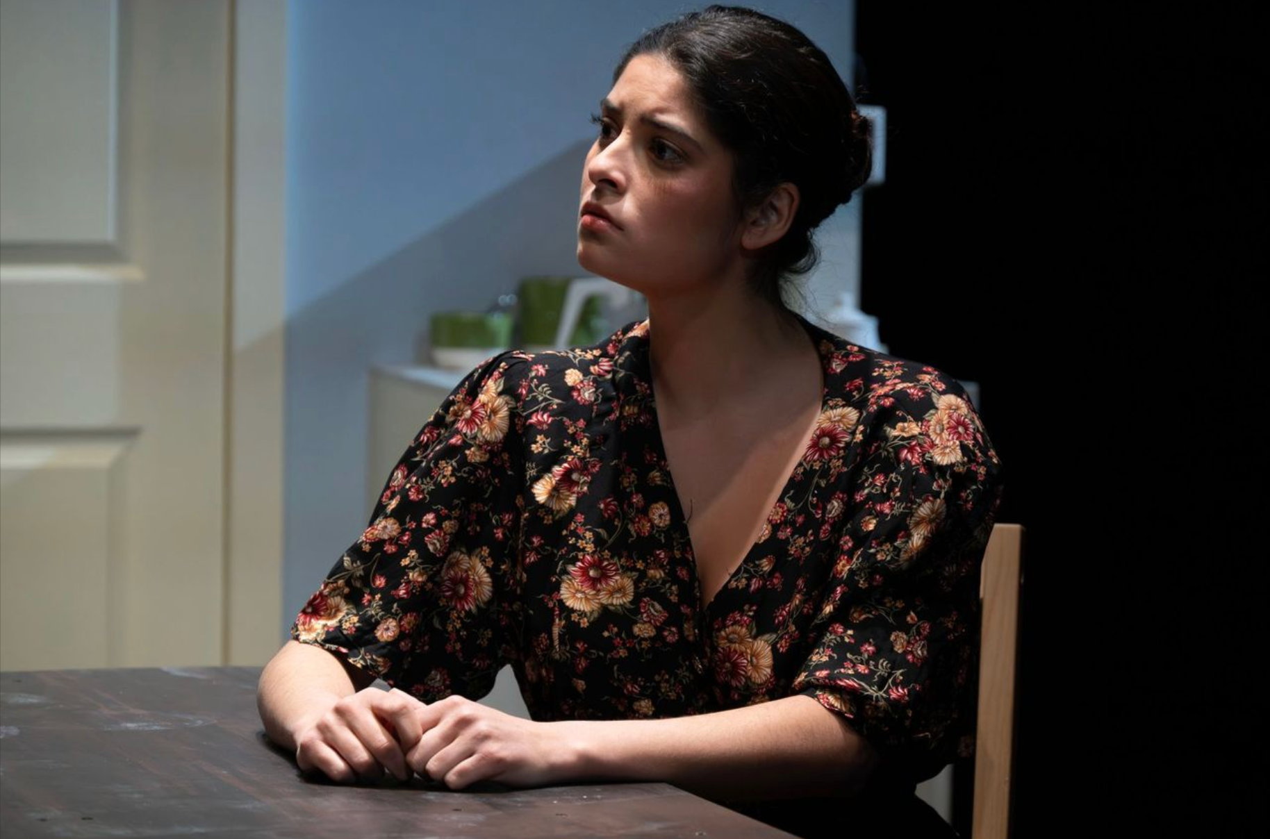 A woman with dark hair styled back, wearing a black floral blouse, sitting at a table with clasped hands, appears thoughtful or concerned, in a dimly lit room with a dark background.