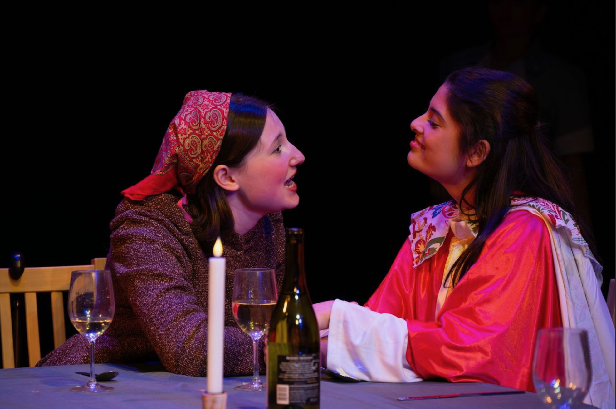 Two women are sitting at a dinner table, smiling and looking at each other. The woman on the left has brown hair, wears a headscarf, and a brown jacket. The woman on the right has dark hair pulled back, wears a colorful scarf and a bright pink jacket. The table has wine glasses, a candle, and a bottle of wine.